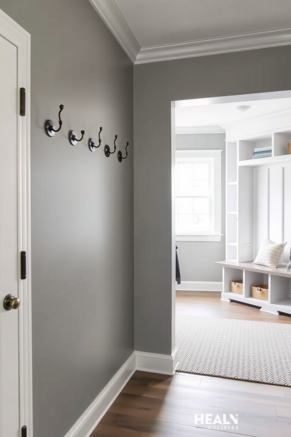 A stylish gray entryway features decorative wall hooks arranged neatly along the wall, providing both function and aesthetic appeal. The floor is adorned with a textured area rug that complements the soft gray tones of the walls. The mudroom showcases a practical design with built-in benches and storage cubbies, seamlessly blending style and utility. Natural light filters through a nearby window, illuminating the space and highlighting the organized, clutter-free environment.