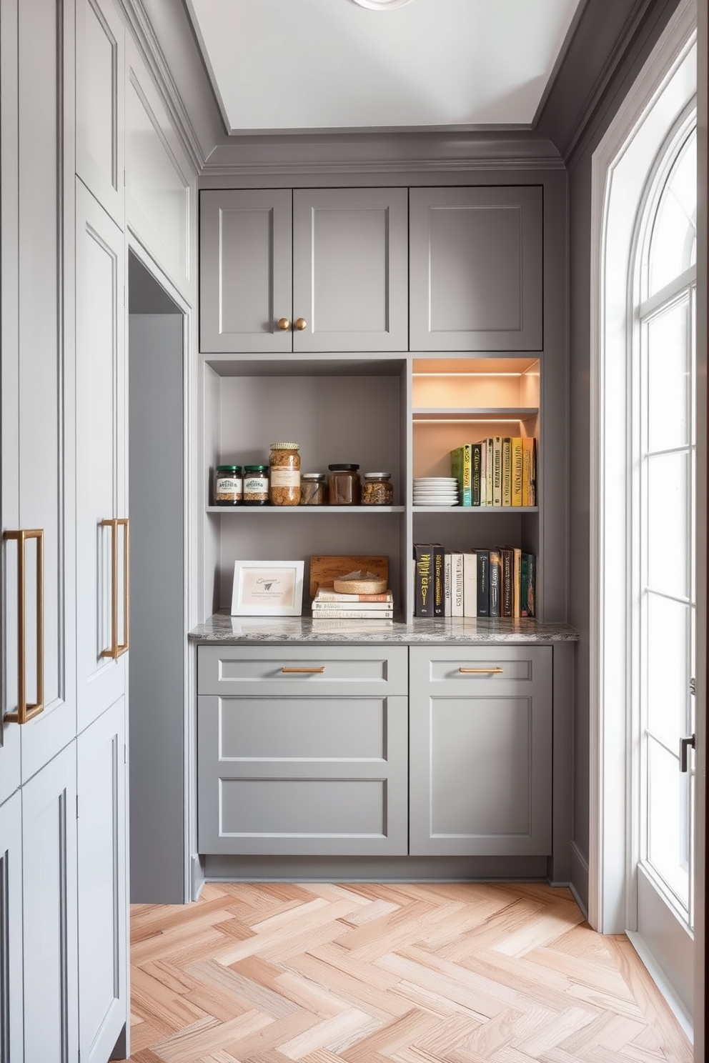 A sophisticated gray pantry with crown molding. The cabinetry features sleek gray doors with elegant hardware, and open shelving displays neatly arranged jars and cookbooks. The walls are painted in a soft gray hue that complements the cabinetry, while the floor showcases a stylish herringbone pattern in light wood. A large window allows natural light to flood the space, highlighting the pantry's organized and inviting atmosphere.