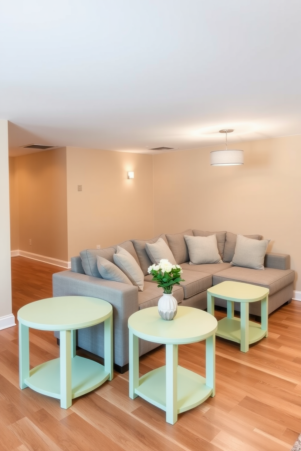 Pastel green side tables add a touch of subtle elegance to the basement space. The walls are painted in a soft beige, complemented by a cozy sectional sofa adorned with light gray cushions. The flooring features a warm wood finish that enhances the inviting atmosphere. Ambient lighting fixtures hang from the ceiling, casting a gentle glow over the room's decor.