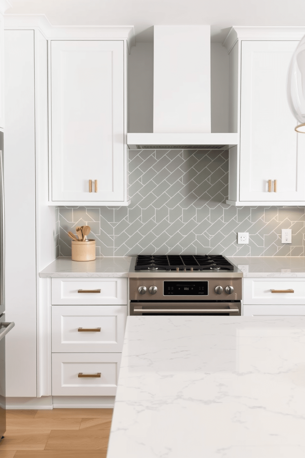 A modern kitchen featuring muted green tiles arranged in a herringbone pattern on the backsplash. The cabinetry is a sleek white with brass hardware, complemented by a spacious island topped with a light marble surface.