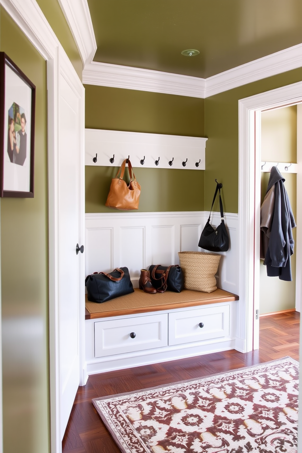 A welcoming mudroom featuring olive green paint on the walls complemented by elegant white wainscoting. The space includes a built-in bench with storage underneath, surrounded by hooks for coats and bags, and a patterned area rug on the floor.