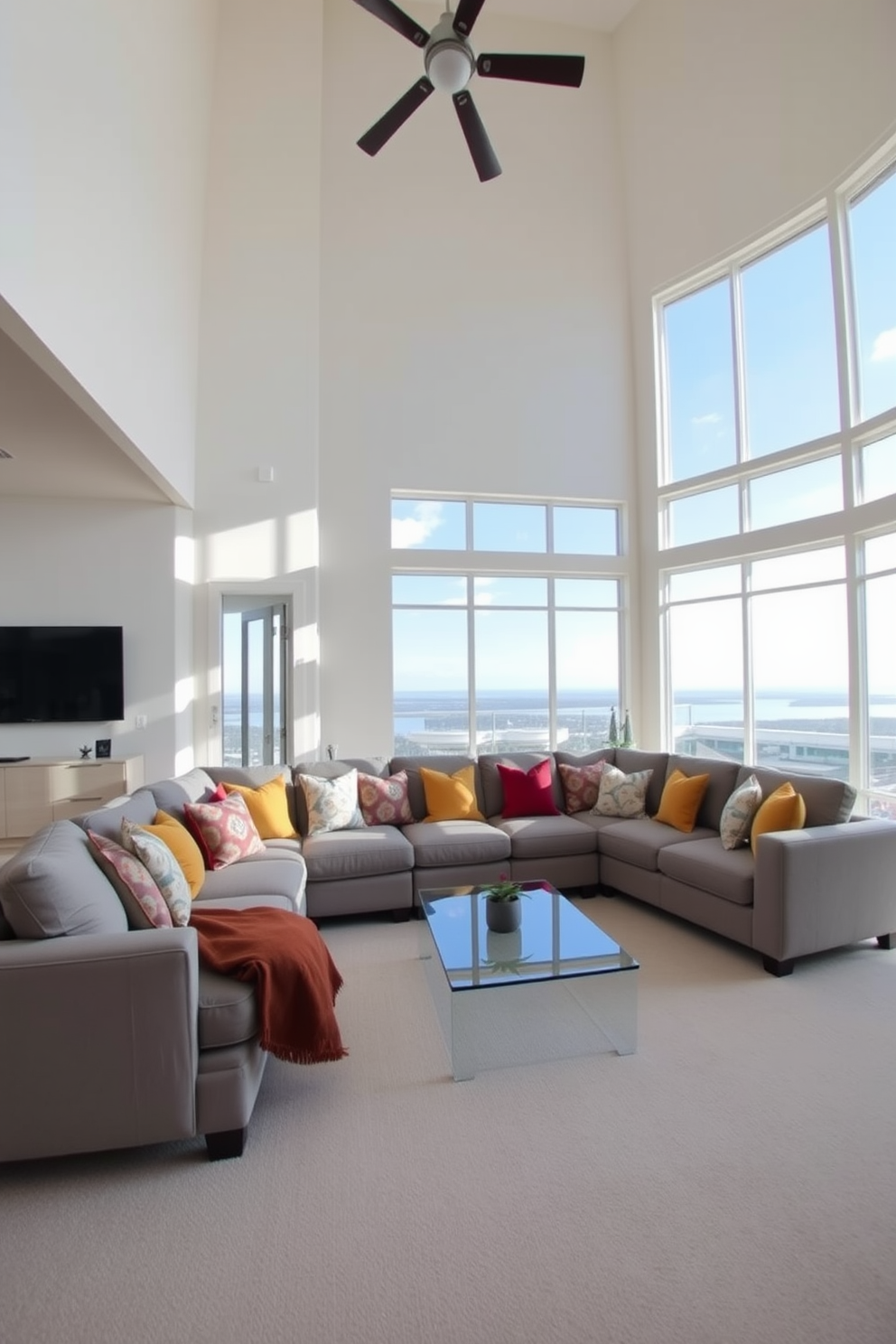 A spacious living room with high ceilings featuring a large sectional sofa in a soft gray fabric. The sofa is adorned with an array of colorful throw pillows, and a sleek glass coffee table sits in front of it. The walls are painted in a warm white tone, creating an inviting atmosphere. Large windows allow natural light to flood the room, enhancing the open feel and showcasing a beautiful view outside.