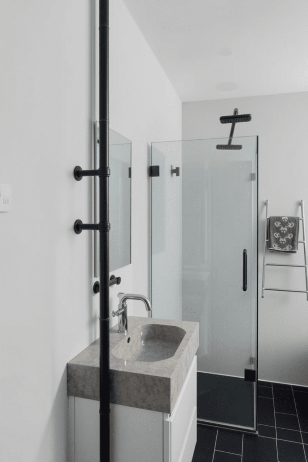 A minimalist bathroom featuring simple lines and geometric shapes. The space includes a concrete sink with a sleek stainless steel faucet and a frameless glass shower enclosure. Exposed pipes and a metal ladder towel rack add to the industrial aesthetic. The walls are painted in a soft gray, complemented by a dark tile floor for contrast.