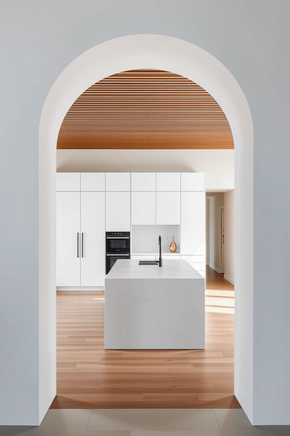 A sleek modern arch frames the entrance to a minimalist kitchen. The arch features clean lines and a smooth finish, complementing the overall contemporary aesthetic. Inside, the kitchen boasts streamlined cabinetry in a matte white finish. A large island with a subtle marble countertop serves as the focal point, enhancing the open and airy feel of the space.