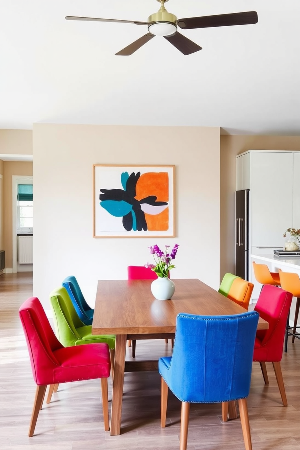 A modern kitchen dining room featuring neutral tones complemented by vibrant color accents. The space includes a sleek wooden dining table surrounded by colorful upholstered chairs that add a playful touch. The walls are painted in a soft beige, while the cabinetry showcases a clean white finish. A bold piece of artwork hangs above the table, bringing energy and personality to the room.