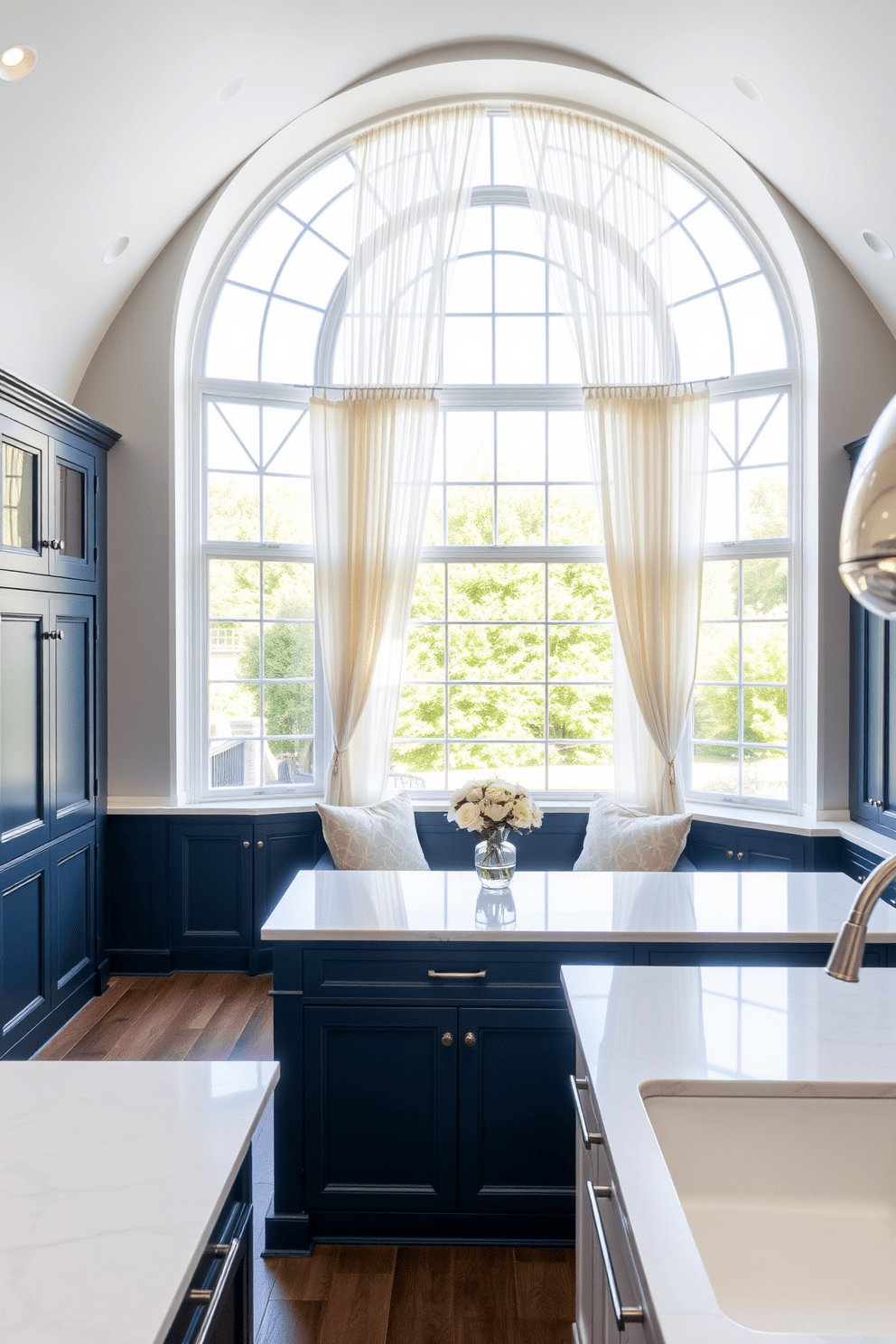 A stunning kitchen design featuring large arched windows that flood the space with natural light. The windows are adorned with soft, flowing curtains that complement the overall color scheme of the kitchen. The cabinetry is a rich navy blue, contrasting beautifully with the white marble countertops. A cozy breakfast nook is nestled beneath the windows, creating an inviting space for morning coffee.