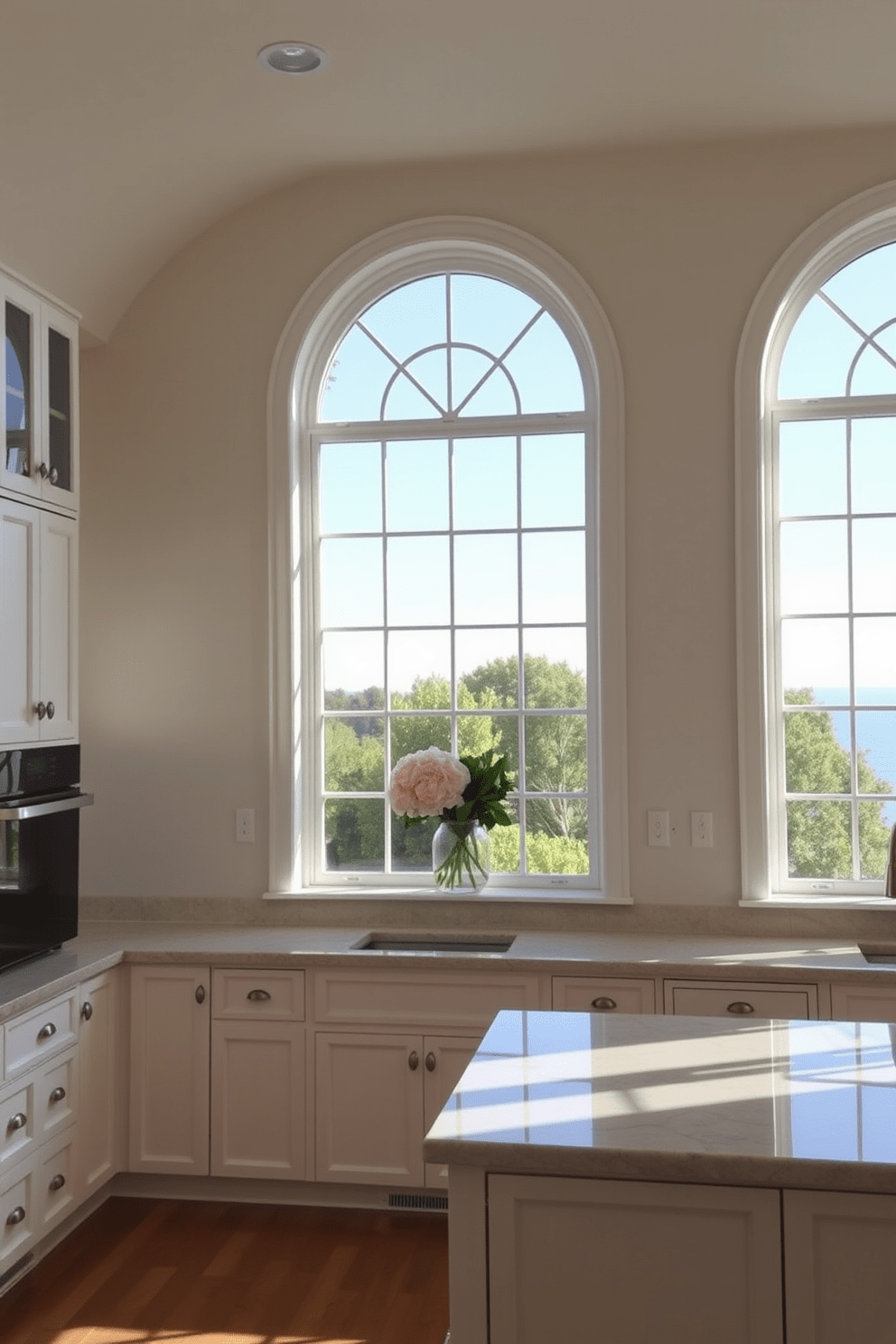 A bright and airy kitchen featuring arched top windows that gracefully frame the view. The soft curves of the windows create a harmonious focal point, allowing natural light to flood the space. The kitchen is designed with a modern aesthetic, showcasing sleek cabinetry in a soft white finish. A spacious island with a marble countertop complements the elegant window design, enhancing the overall warmth and charm of the room.