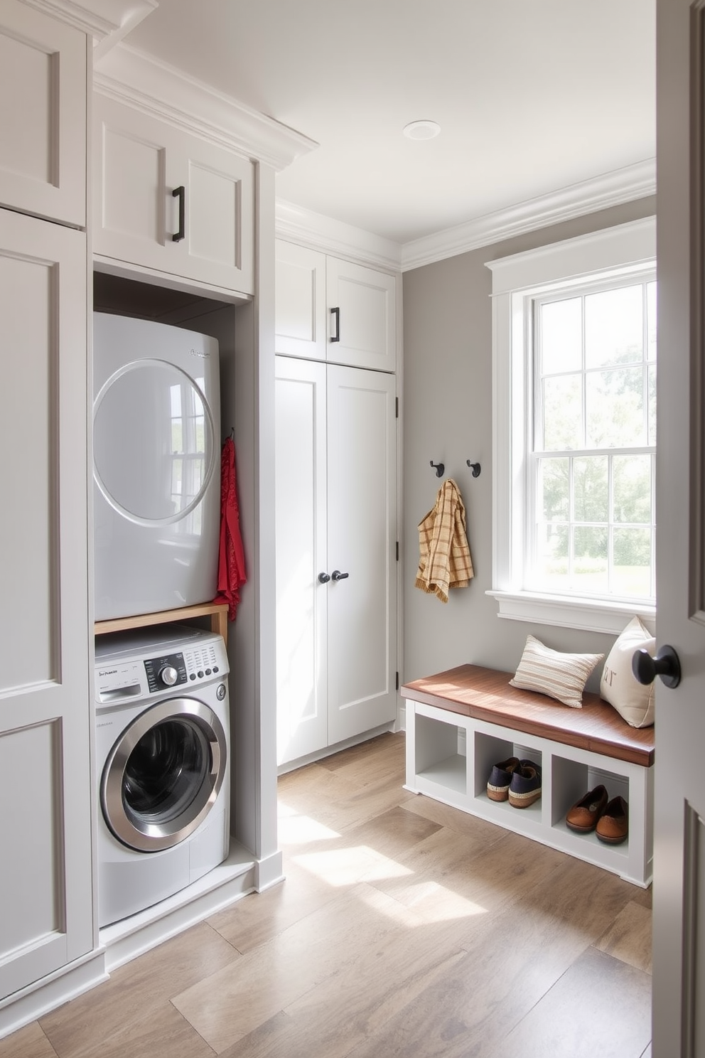 A spacious mudroom features an integrated laundry area with a stacked washer and dryer tucked neatly into custom cabinetry. The room is designed with durable, easy-to-clean flooring and ample storage solutions, including hooks for coats and baskets for shoes. Natural light floods the space through a large window, illuminating the soft gray walls and white trim. A stylish bench with cushions provides a comfortable spot to sit while removing shoes, enhancing the functionality and aesthetics of the room.