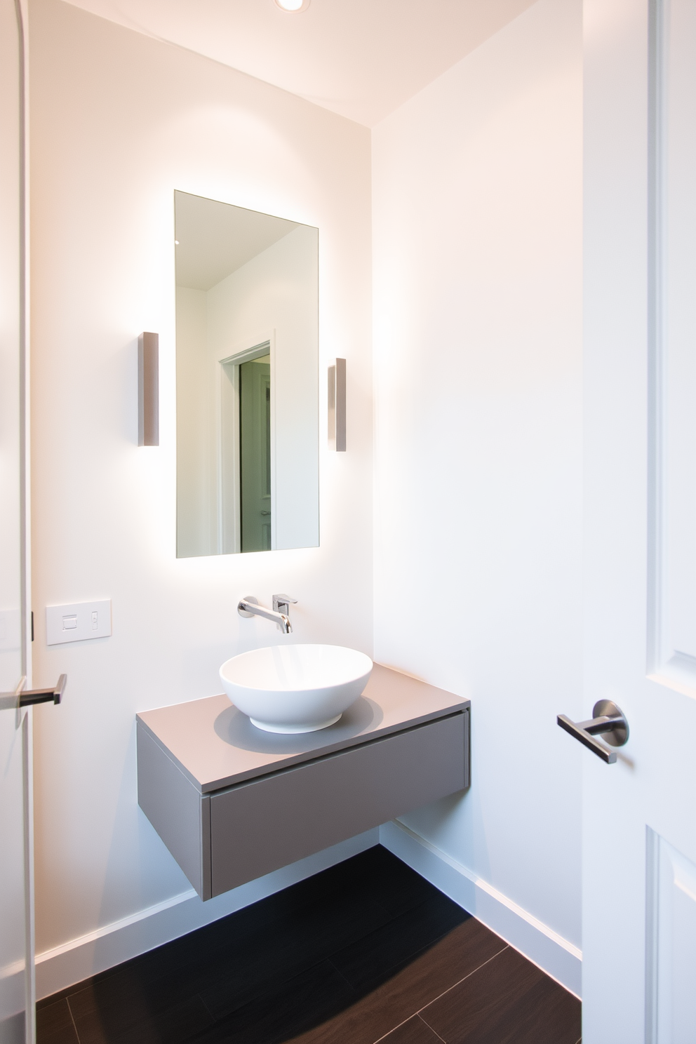 A large powder room features sleek modern fixtures that enhance its contemporary style. The space is adorned with a floating vanity that boasts a minimalist design, complemented by a stylish vessel sink and a wall-mounted faucet. The walls are painted in a crisp white, creating a bright and airy atmosphere. Accent lighting highlights the elegant details, while a large mirror reflects the sophisticated elements of the room.