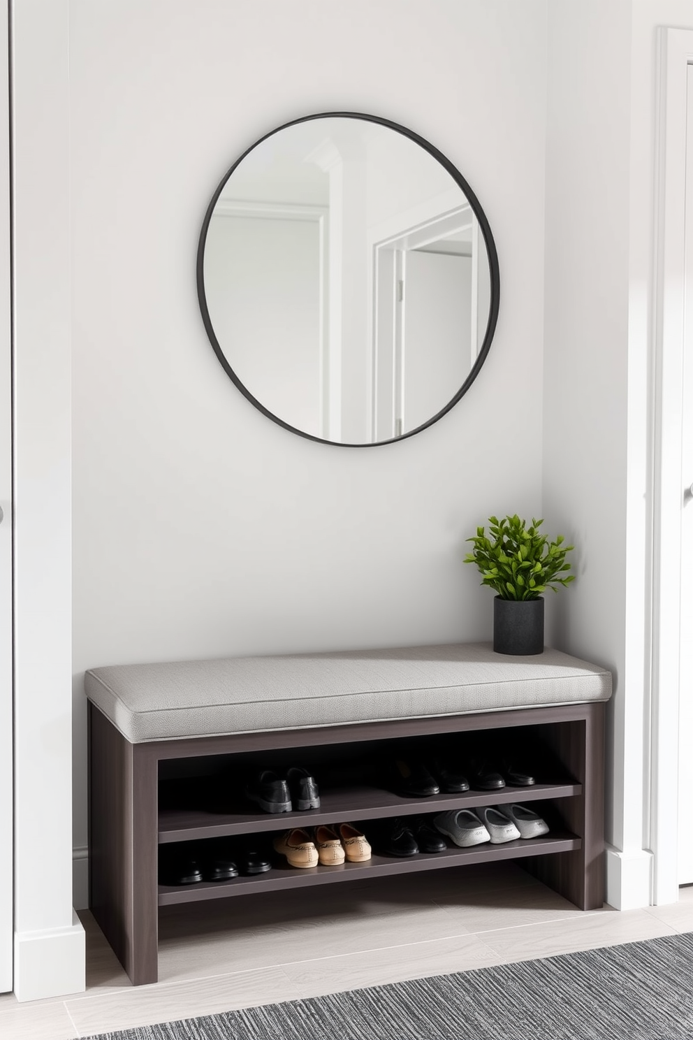 A contemporary entryway featuring a sleek shoe storage unit seamlessly integrated under a stylish bench. The bench is upholstered in a soft gray fabric, providing comfort while maintaining a minimalist aesthetic. The walls are painted in a crisp white, enhancing the sense of space, while a large round mirror hangs above the bench, reflecting light and creating depth. A small potted plant sits beside the bench, adding a touch of greenery to the modern design.