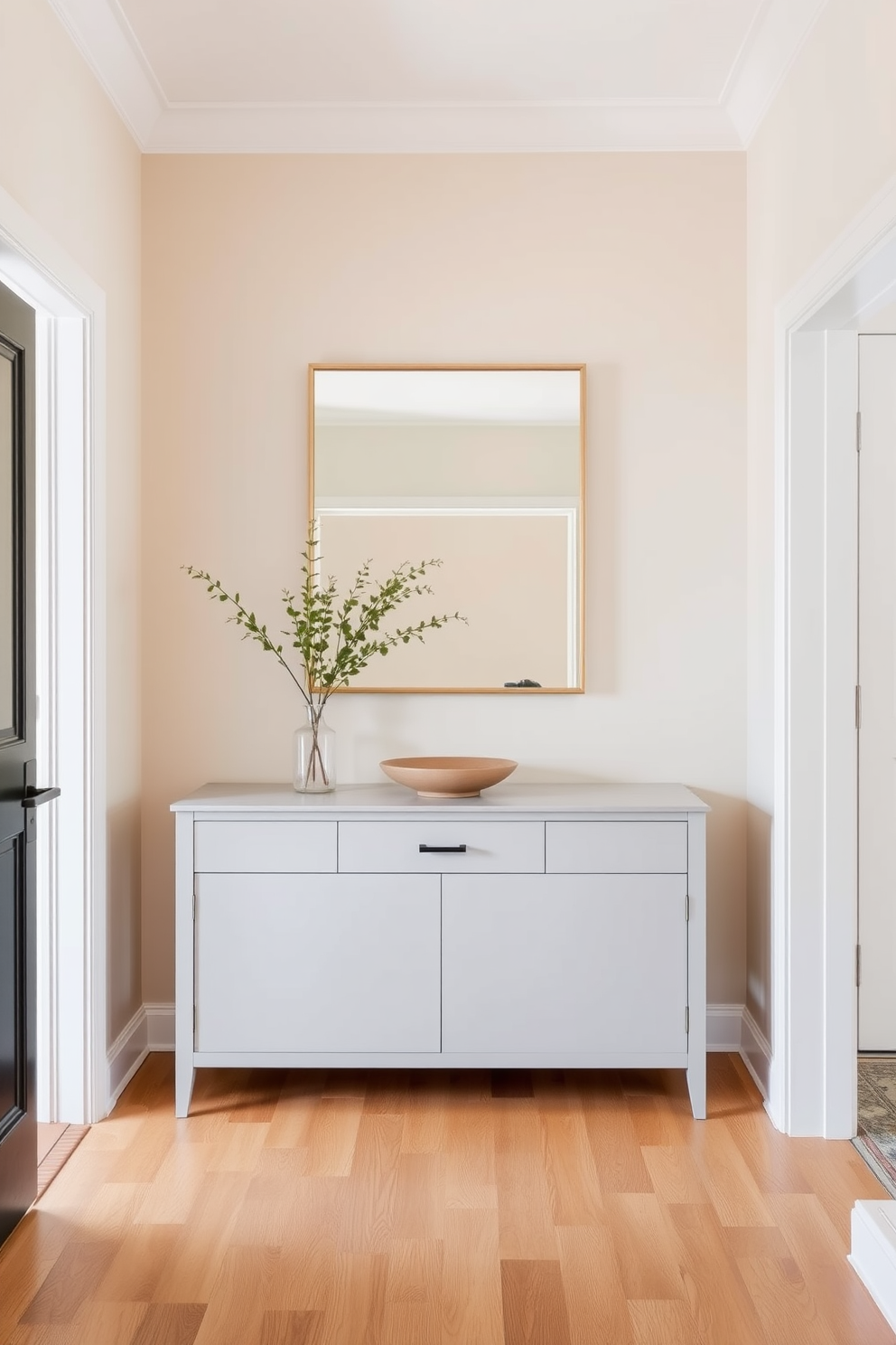 A long foyer with a neutral color palette creates a calming and inviting atmosphere. Soft beige walls complement a light gray console table adorned with a simple decorative bowl and a small potted plant. The floor features wide plank hardwood in a warm tone, enhancing the serene vibe of the space. A large mirror with a minimalist frame hangs above the console, reflecting natural light and making the foyer feel more spacious.