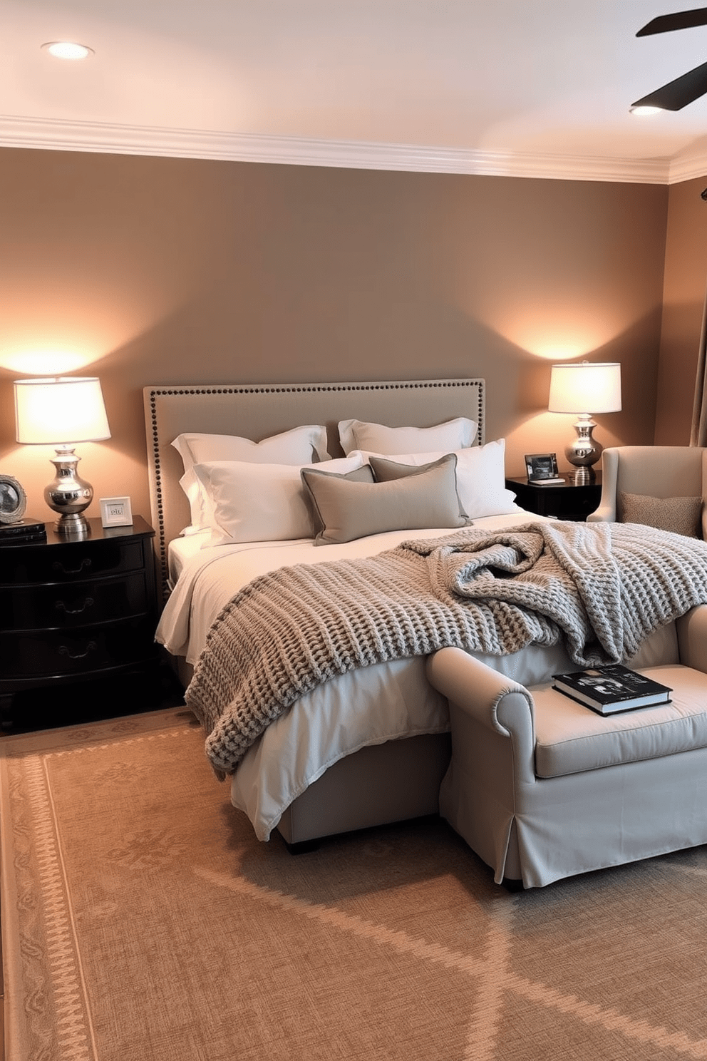 Create a cozy atmosphere with warm lighting in a master bedroom. The room features a plush king-sized bed dressed in soft, neutral linens with an oversized knitted throw draped across the foot. On either side of the bed, there are matching bedside tables with elegant lamps that emit a soft glow. A comfortable armchair is placed in the corner, accompanied by a small side table and a stack of books for a relaxing reading nook. The walls are painted a calming taupe color, enhancing the warmth of the space. A large area rug with a subtle pattern anchors the room, adding texture and comfort underfoot.