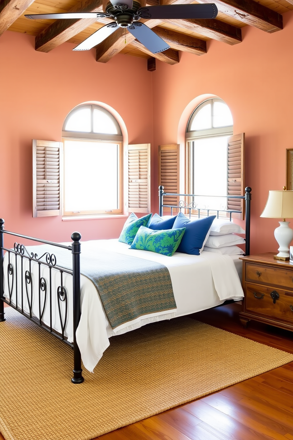 A serene Mediterranean bedroom featuring rustic wooden shutters on the windows. The walls are painted in a soft terracotta hue, complemented by a woven jute rug on the warm wooden floor. A large wrought iron bed with crisp white linens takes center stage. Decorative pillows in vibrant shades of blue and green add a pop of color, while a vintage wooden chest serves as a bedside table.