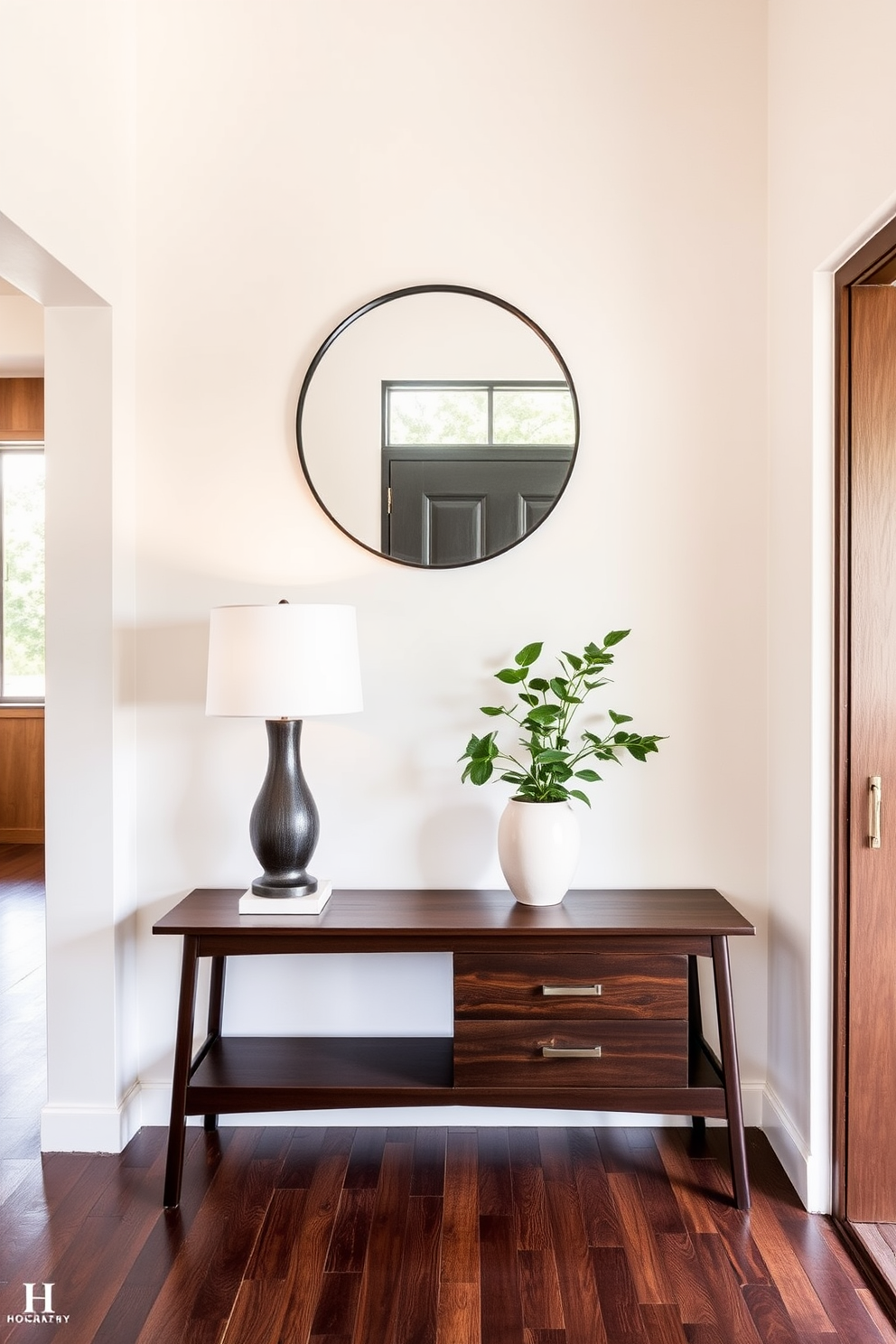 A Mid Century Modern foyer featuring a striking contrast between light and dark finishes. The walls are painted in a soft cream color, while a dark wood console table stands against them, topped with a sculptural lamp and a vibrant potted plant. The floor showcases rich walnut planks that complement the dark tones of the furniture. A large round mirror with a thin black frame hangs above the console, reflecting natural light that filters in through a nearby window.