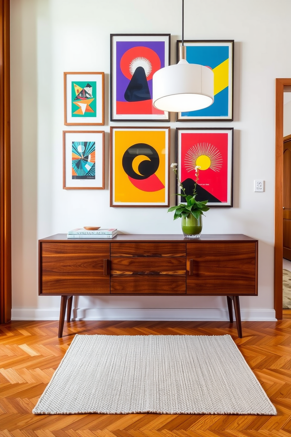 A stylish foyer featuring a sleek console table made of rich walnut wood. Above the table, a collection of framed art prints with bold graphics in vibrant colors creates an eye-catching gallery wall. The flooring is a warm herringbone pattern, complemented by a plush area rug in neutral tones. A modern pendant light hangs from the ceiling, casting a soft glow over the space and enhancing the Mid Century Modern aesthetic.