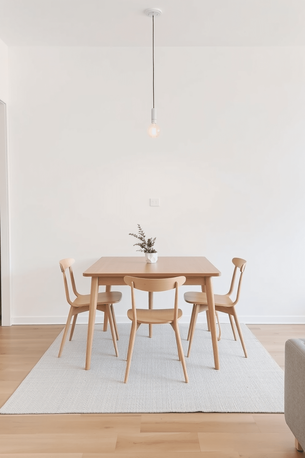 A simple dining table made of light wood sits in the center of the room surrounded by four matching minimalist chairs. The walls are painted in a soft white hue, and a single pendant light hangs above the table, casting a warm glow over the space. A small potted plant rests on the table, adding a touch of greenery to the minimalist aesthetic. The floor is adorned with a light gray rug that complements the overall clean and airy feel of the apartment.