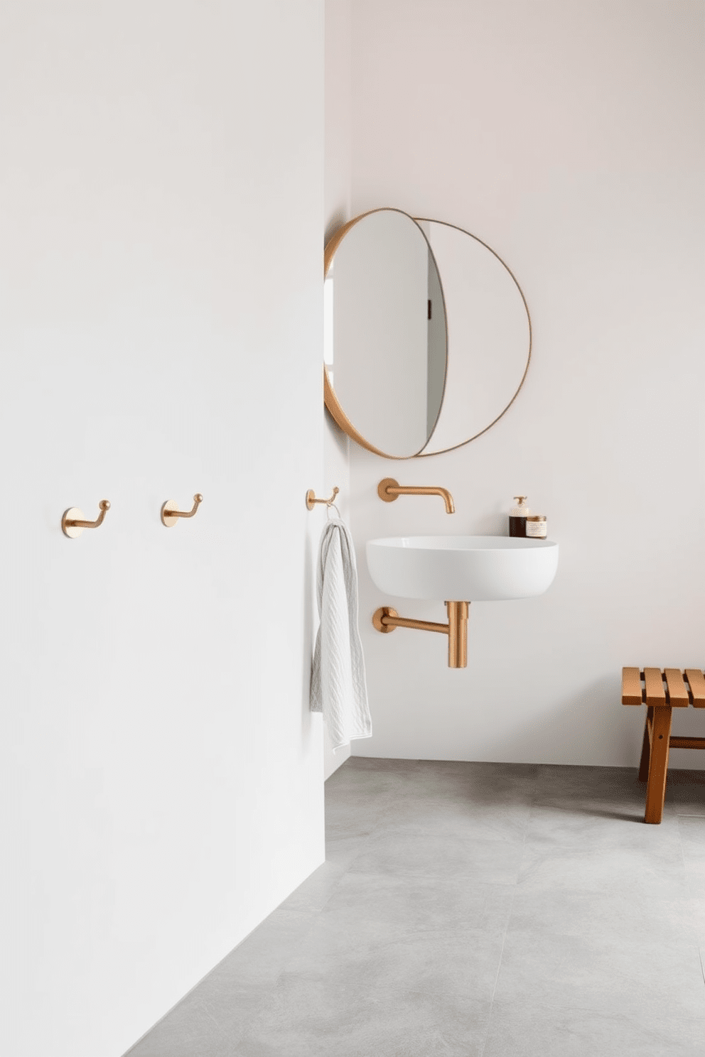 A minimalist bathroom design featuring decorative yet functional towel hooks. The hooks are crafted from brushed brass and are mounted on a smooth white wall, providing an elegant touch while maintaining a clean aesthetic. The floor is adorned with large-format gray tiles, and a simple wooden bench sits against one wall for added functionality. A large round mirror above the sink reflects natural light, enhancing the serene atmosphere of the space.