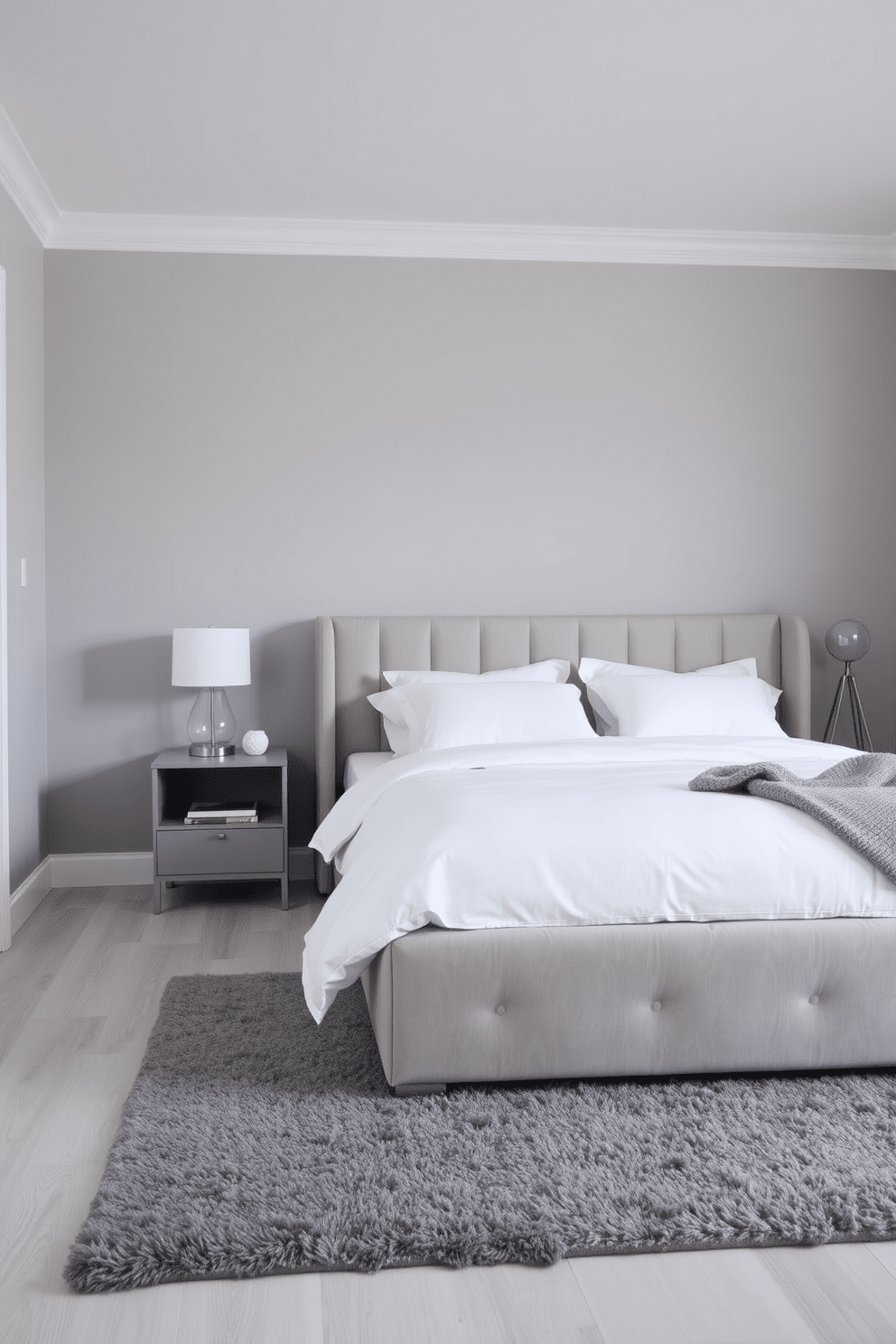 A serene modern bedroom featuring a monochromatic color scheme. The walls are painted in soft gray, complemented by a plush gray upholstered bed with crisp white bedding and matching pillows. A sleek nightstand in a matching gray tone sits beside the bed, adorned with a minimalist lamp. The flooring is a light gray hardwood, and a cozy area rug in a darker shade of gray adds warmth to the space.