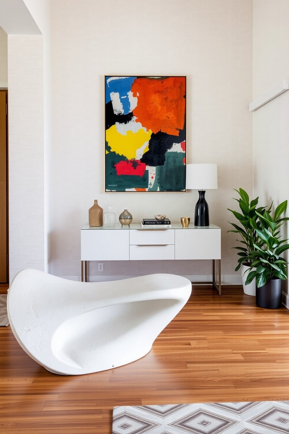 A modern entryway features a striking sculptural bench made of molded concrete, positioned against a backdrop of textured wallpaper in soft neutral tones. Above the bench, a large abstract artwork in vibrant colors draws the eye, while a sleek console table with minimalist design holds decorative items and a stylish lamp. The flooring is a combination of polished hardwood and geometric patterned tiles, creating visual interest and warmth. Potted greenery flanks the entryway, adding a touch of nature and freshness to the space.