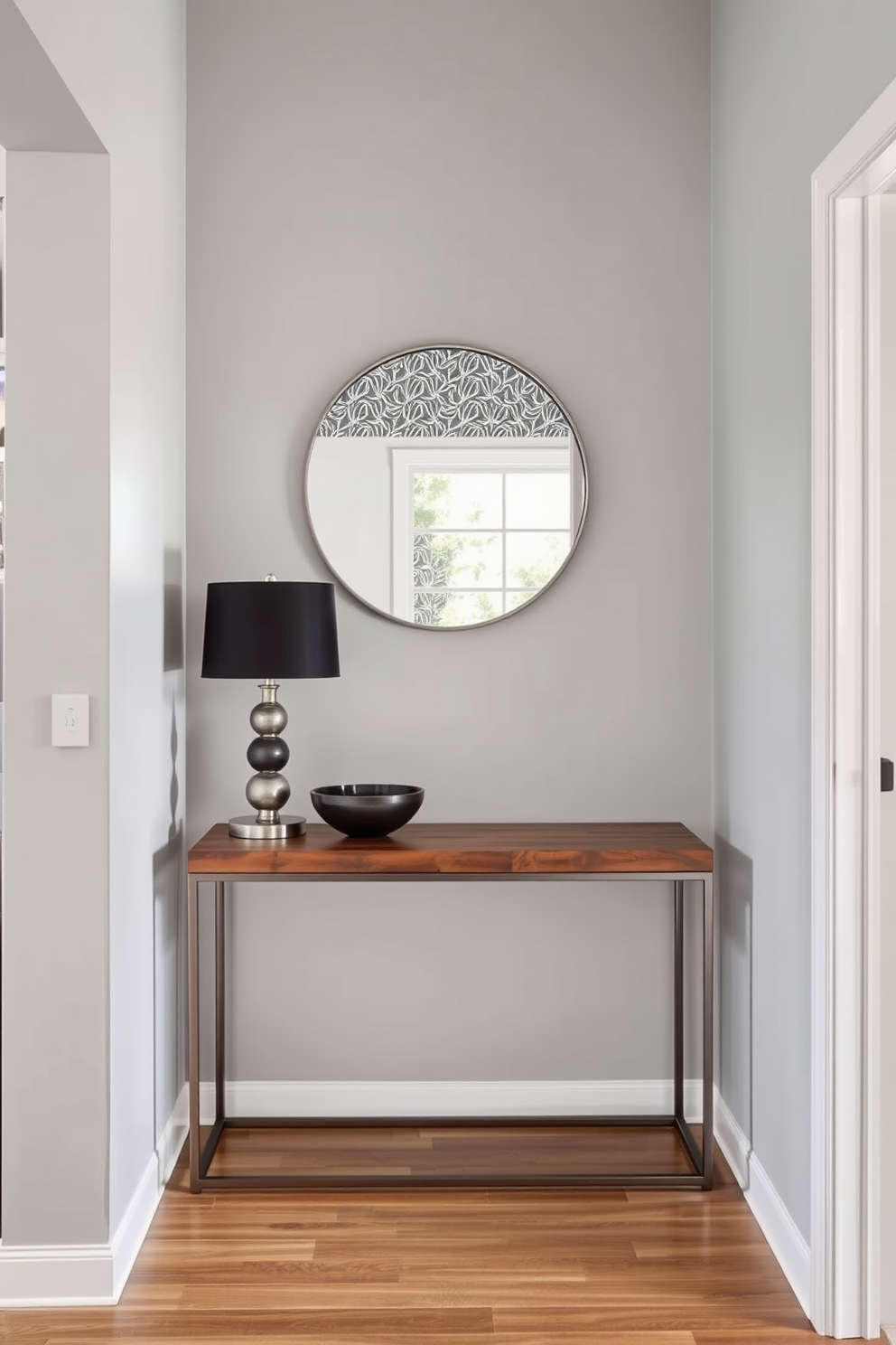 A modern foyer design featuring a striking combination of wood and metal elements. The entrance showcases a sleek metal console table with a rich wooden top, adorned with a stylish lamp and a decorative bowl. The walls are painted in a soft gray tone, complemented by a bold geometric wallpaper on one accent wall. A large round mirror with a metal frame hangs above the console, reflecting natural light from a nearby window.