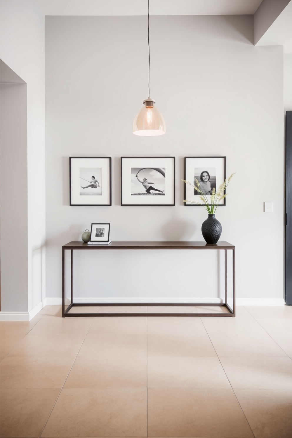 A modern foyer featuring a sleek console table against a light gray wall. Above the table, a series of framed photographs in minimalist frames add a personal touch to the space. The floor is adorned with large format tiles in a soft beige tone, creating a warm and inviting entrance. A contemporary pendant light hangs from the ceiling, illuminating the area with a soft glow.