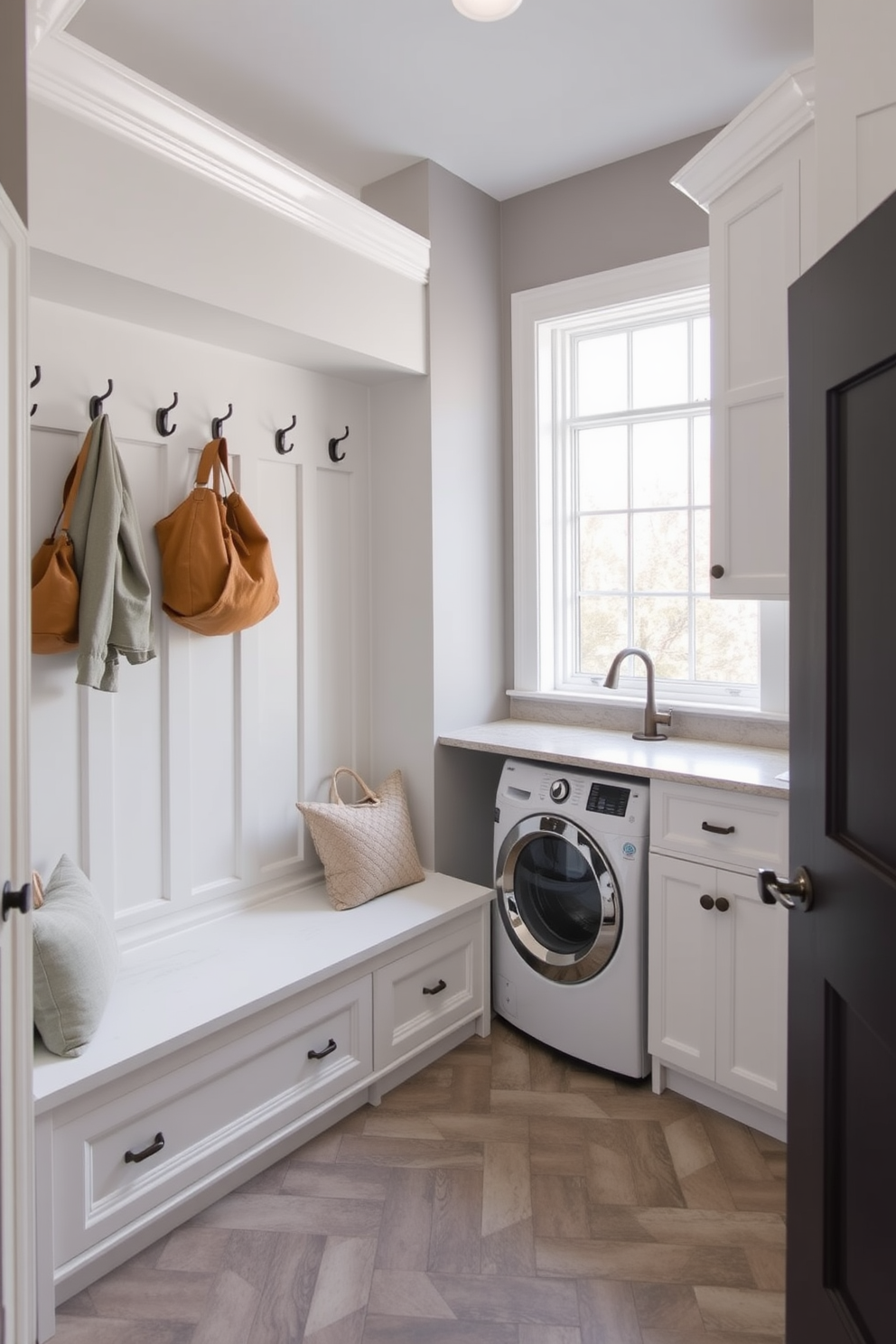 A functional mudroom-laundry combo space features built-in storage benches with hooks above for coats and bags. The laundry area includes stacked machines with a countertop for folding clothes and a large window that brings in natural light. The walls are painted in a soft gray hue, complemented by white cabinetry for a clean look. A durable tile floor in a herringbone pattern adds texture and is easy to maintain.