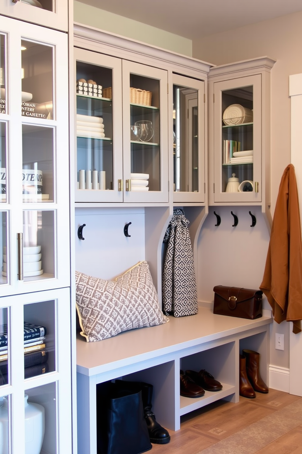 A modern mudroom features glass-front cabinets that showcase neatly organized items and stylish decor. The space includes a bench with built-in storage, hooks for coats, and a neutral color palette that enhances the overall design.