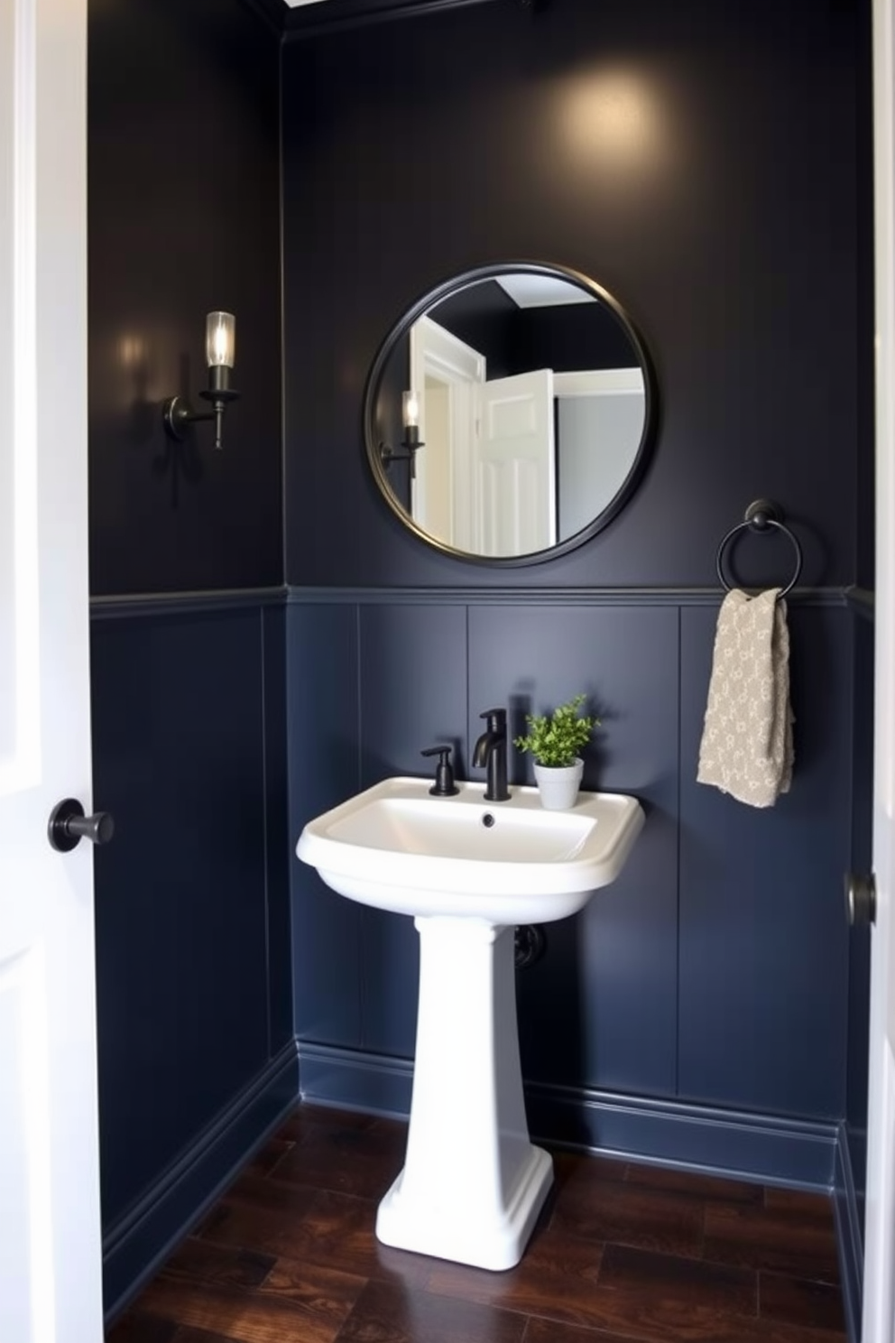 A moody powder room with dark wood flooring that creates a rich foundation. The walls are painted in a deep navy blue, complemented by sleek black fixtures and a stylish pedestal sink. A large round mirror with a black frame hangs above the sink, reflecting the soft glow of vintage-style wall sconces. A small potted plant sits on the countertop, adding a touch of greenery to the sophisticated space.