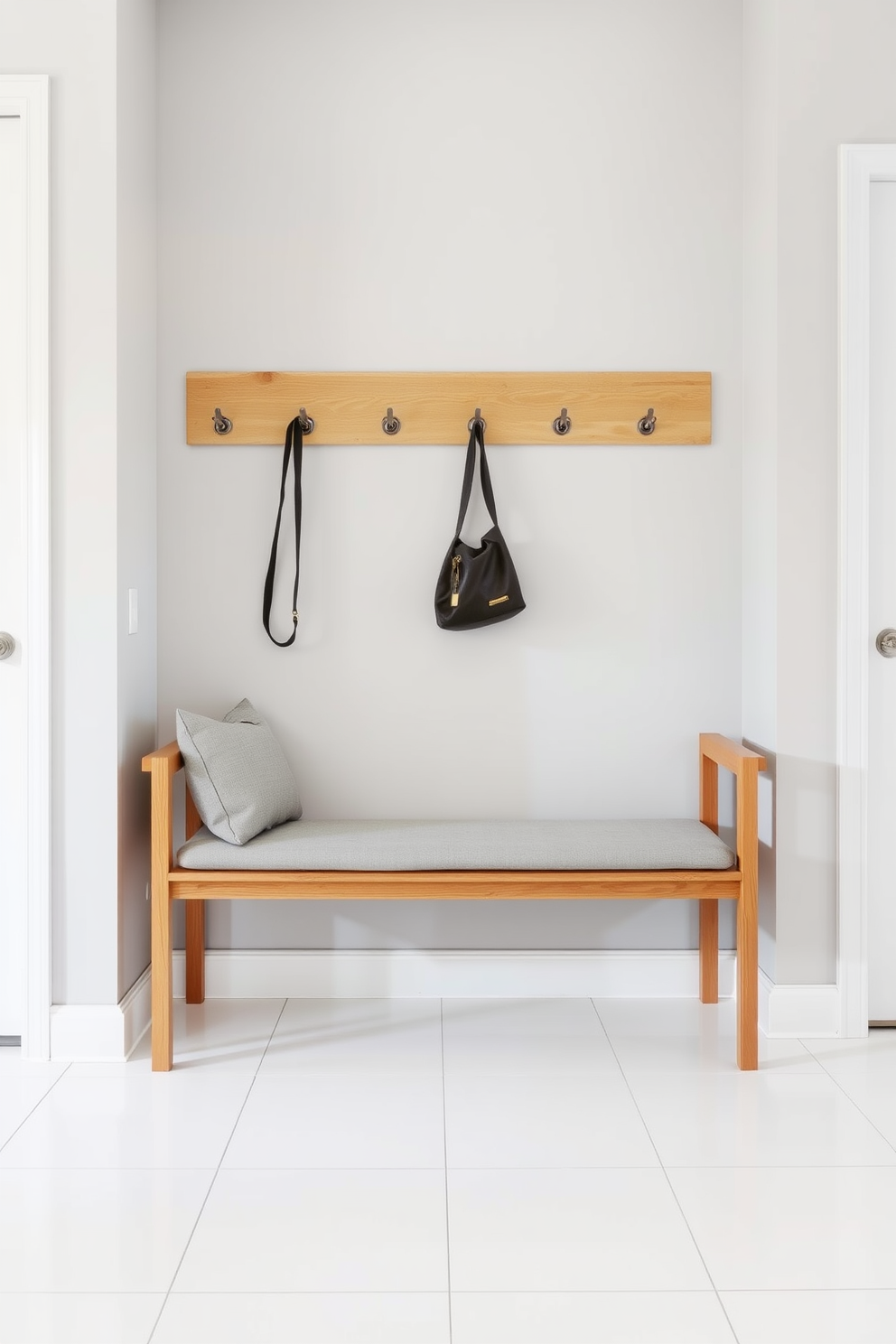 A modern minimalist bench with sleek lines is positioned against a light gray wall. The bench features a natural wood finish and is complemented by a soft gray cushion for added comfort. In the mudroom, the bench is paired with hooks for hanging coats and bags. The floor is adorned with large white tiles, creating a clean and airy atmosphere.