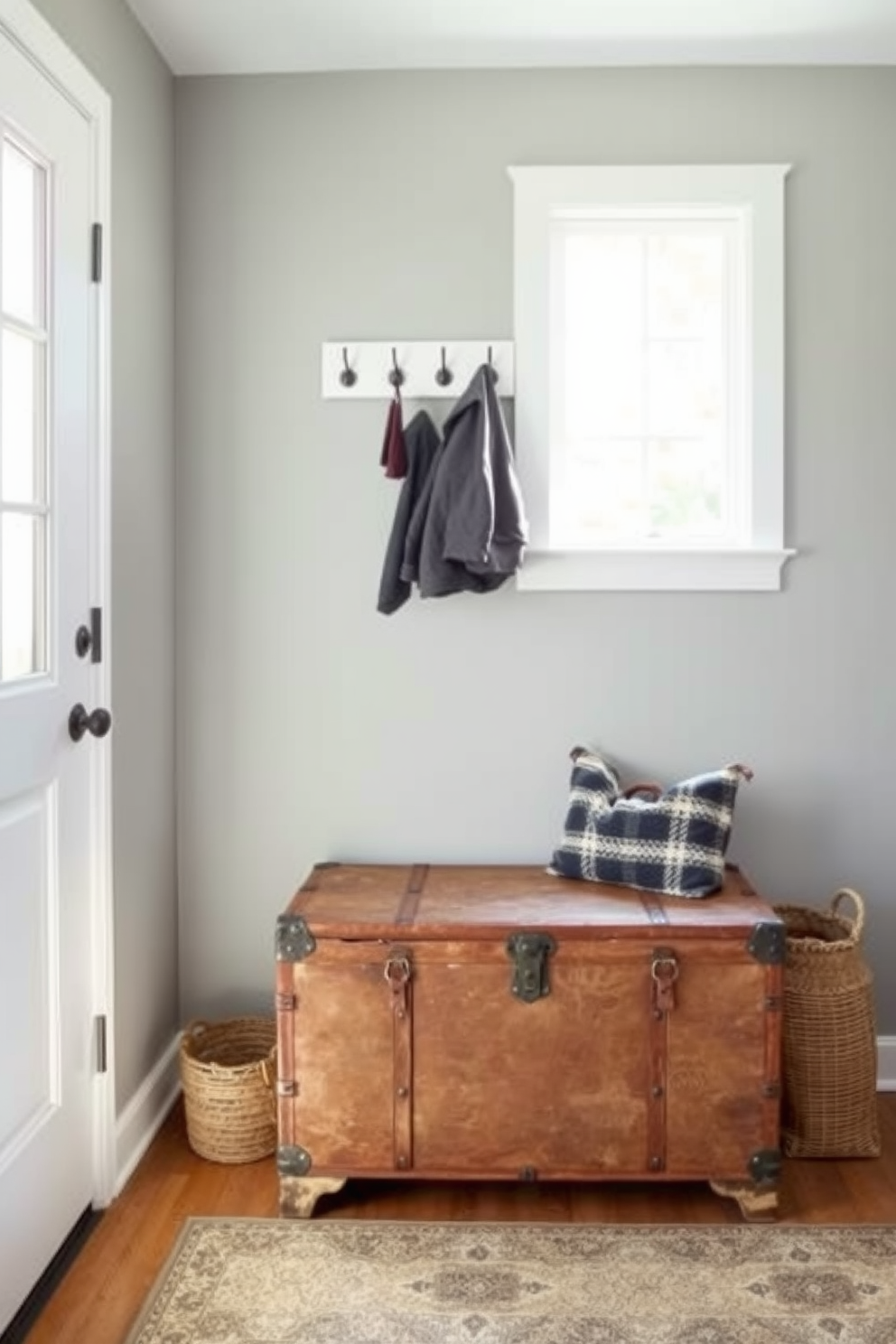 A cozy mudroom features a vintage trunk repurposed as a seat, providing both storage and a charming focal point. The walls are painted in a soft gray, and a woven basket sits beside the trunk, adding warmth and texture to the space. Natural light floods in through a nearby window, illuminating a collection of hooks above the trunk for hanging coats and bags. The floor is adorned with a patterned rug that complements the rustic aesthetic of the vintage trunk.