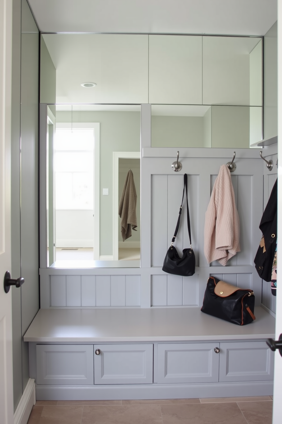 A stylish mudroom featuring a mirrored wall that enhances the sense of space and light. The design includes built-in storage benches with hooks above for coats and bags, all in a soft gray palette.