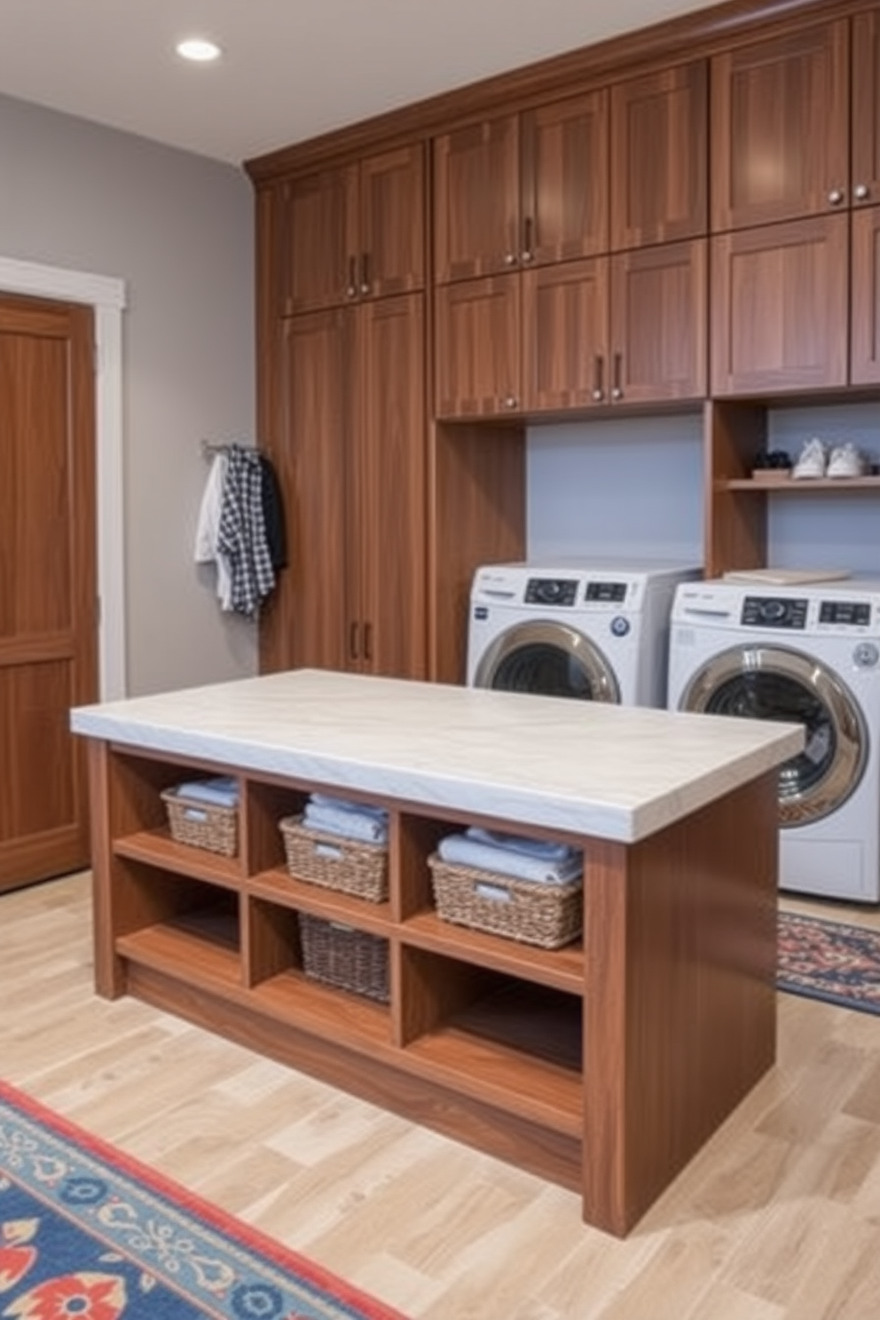 A stylish mudroom island designed for folding clothes features a spacious countertop made of quartz with a subtle veining pattern. Beneath the countertop, open shelves provide ample storage for baskets filled with linens and a built-in bench offers a comfortable spot to sit and remove shoes. The mudroom laundry room design incorporates a functional layout with side-by-side washer and dryer units tucked into cabinetry. Light gray walls complement the rich wood tones of the cabinetry, while a colorful rug adds a pop of personality to the space.