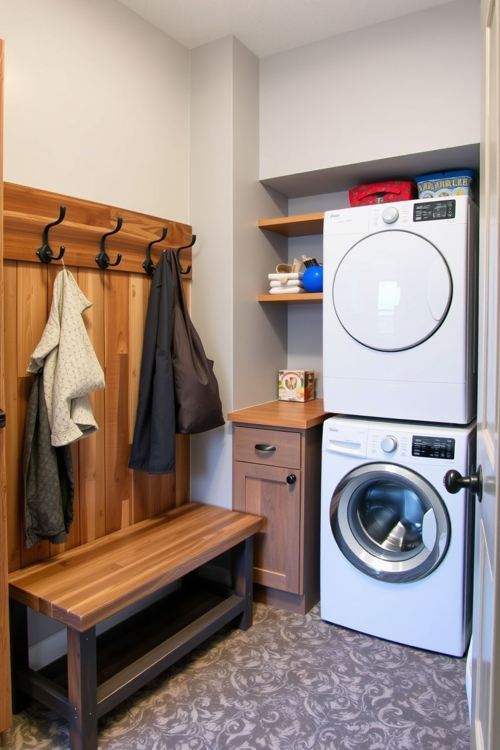 A cozy mudroom features rustic wood accents with a combination of metal fixtures. The space includes a built-in bench made of reclaimed wood, paired with hooks for hanging coats and bags. Adjacent to the bench, a functional laundry area showcases a stacked washer and dryer with a wood countertop above for folding clothes. The walls are painted a soft gray, while the floor is covered in durable, patterned tile that adds character to the room.