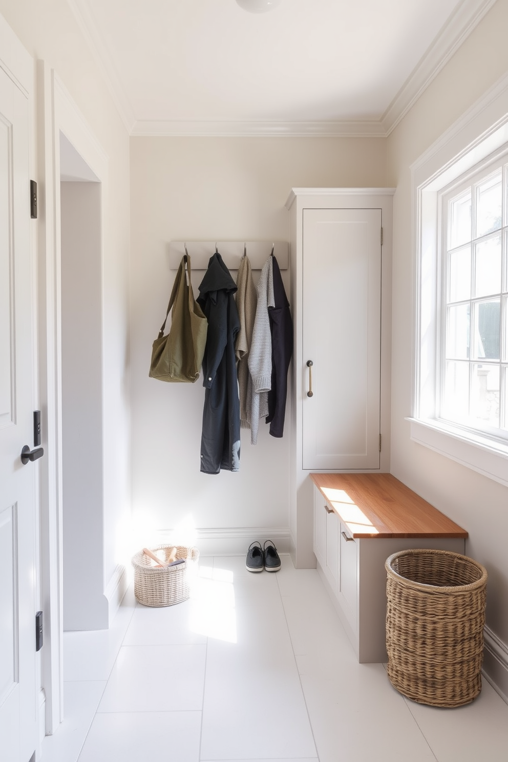 A serene mudroom with a neutral color palette featuring soft beige walls and light gray cabinetry. The floor is adorned with large white tiles, and a wooden bench with storage underneath provides a welcoming touch. To the left, a row of hooks is mounted on the wall for coats and bags, while a woven basket sits on the floor for shoes. Natural light pours in through a nearby window, enhancing the calming atmosphere of the space.