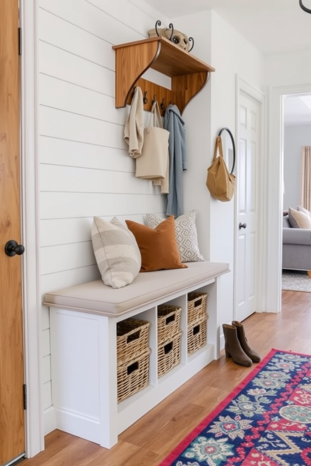 A cozy bench with storage underneath is placed in the mudroom, providing a welcoming area for family members to sit and remove their shoes. The bench is upholstered in soft fabric and features decorative pillows, while the storage underneath is designed with woven baskets for organization. Adjacent to the family room, the mudroom design incorporates a light color palette with shiplap walls and warm wood accents. A stylish coat rack is mounted above the bench, and a vibrant area rug adds a pop of color to the space, creating a seamless transition between the mudroom and the family room.