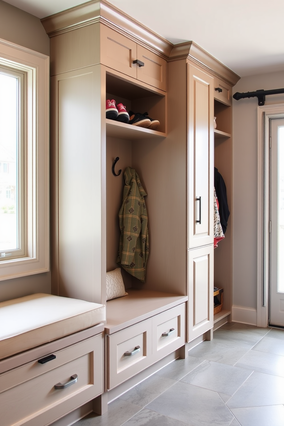 A functional mudroom space near the garage features built-in cabinetry with a mix of open and closed storage. The design includes a bench with cushioned seating, hooks for coats, and a dedicated area for charging devices, all accented with a neutral color palette. The charging station is seamlessly integrated into the cabinetry, providing easy access for devices while maintaining a clutter-free environment. Natural light floods the space through a nearby window, highlighting the durable flooring and stylish organizational elements.