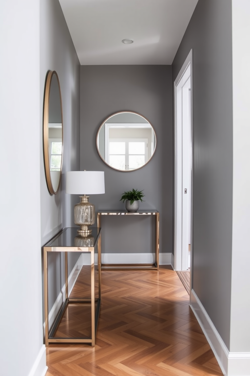 A mirrored entryway that reflects light and creates an illusion of space. The walls are painted in a soft gray, and a sleek console table is placed against one side with a decorative lamp on top. The floor features elegant herringbone wood, enhancing the narrow design. A large round mirror hangs above the console, complemented by a small potted plant for a touch of greenery.