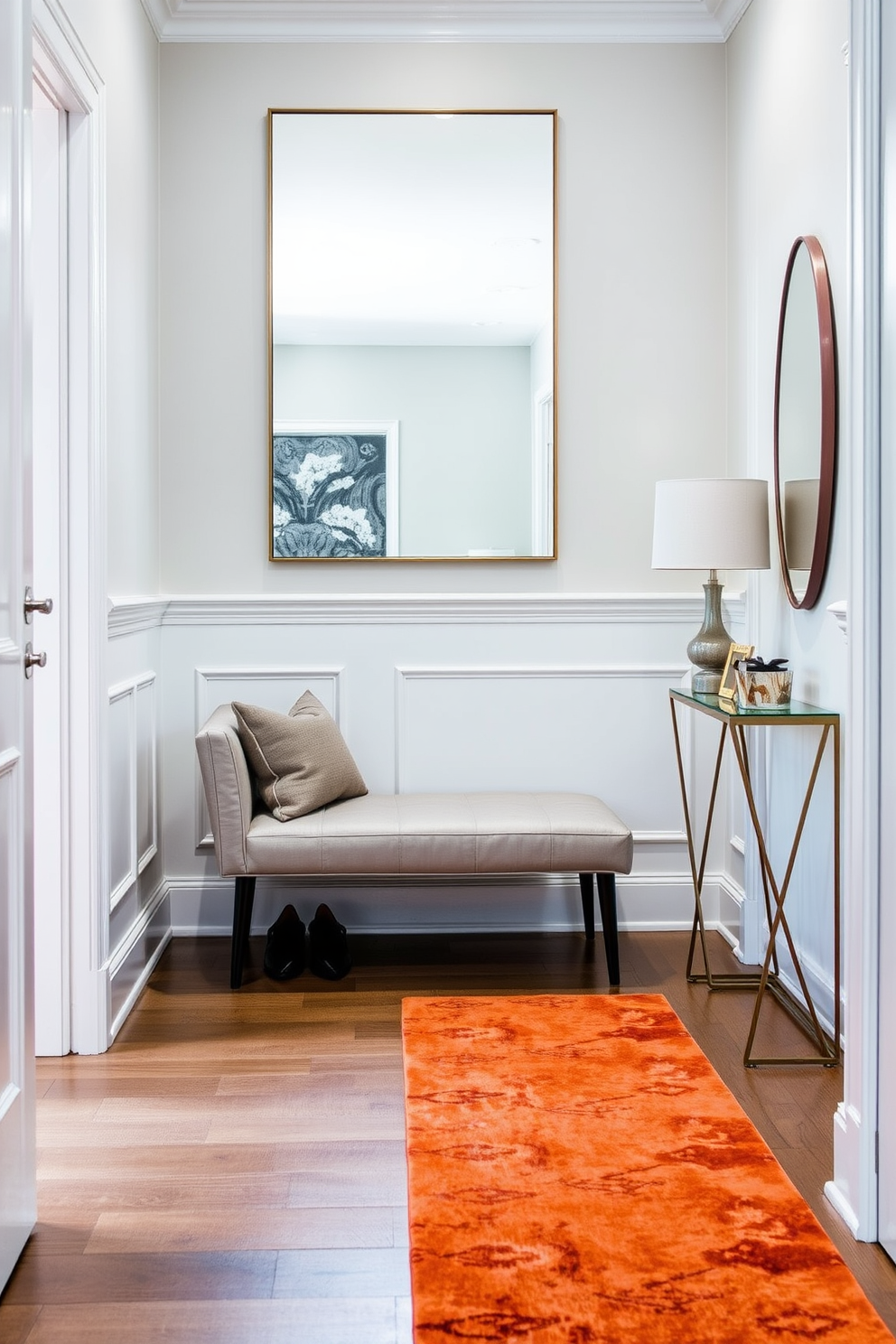 A narrow foyer features a sleek bench upholstered in soft gray fabric, providing a comfortable spot for putting on shoes. The walls are adorned with elegant wainscoting painted in a crisp white, while a large mirror above the bench enhances the sense of space and light. To the side, a small console table holds decorative items and a stylish lamp, adding functionality and charm. A vibrant runner rug in warm tones stretches along the floor, inviting guests into the home with a pop of color.