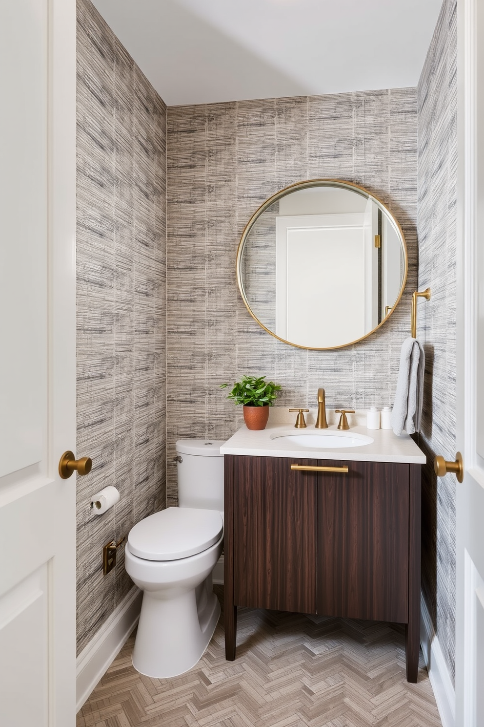 A narrow powder room features layered textures that create visual interest. The walls are adorned with a textured wallpaper, complemented by a sleek, dark wood vanity topped with a light-colored stone surface. A large round mirror hangs above the vanity, framed in brushed brass to add warmth. The floor is covered in a herringbone-patterned tile, while a small potted plant adds a touch of greenery to the space.