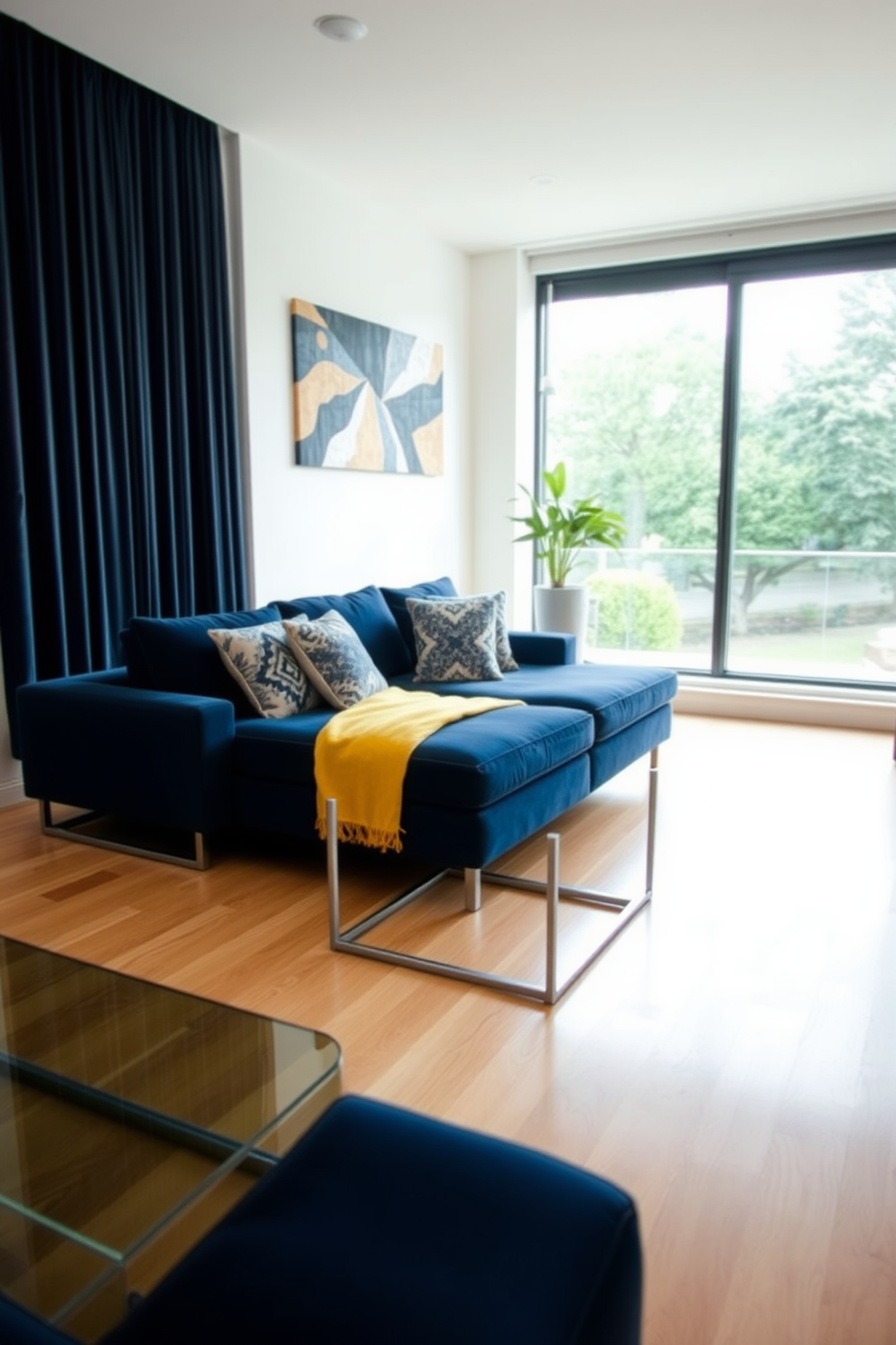 A stylish living room featuring a navy blue couch that serves as the focal point. The light wood flooring complements the rich color of the couch, creating a warm and inviting atmosphere. In the background, a large window allows natural light to flood the space, highlighting decorative throw pillows on the couch. A sleek coffee table made of glass and metal sits in front of the couch, adding a modern touch to the room.