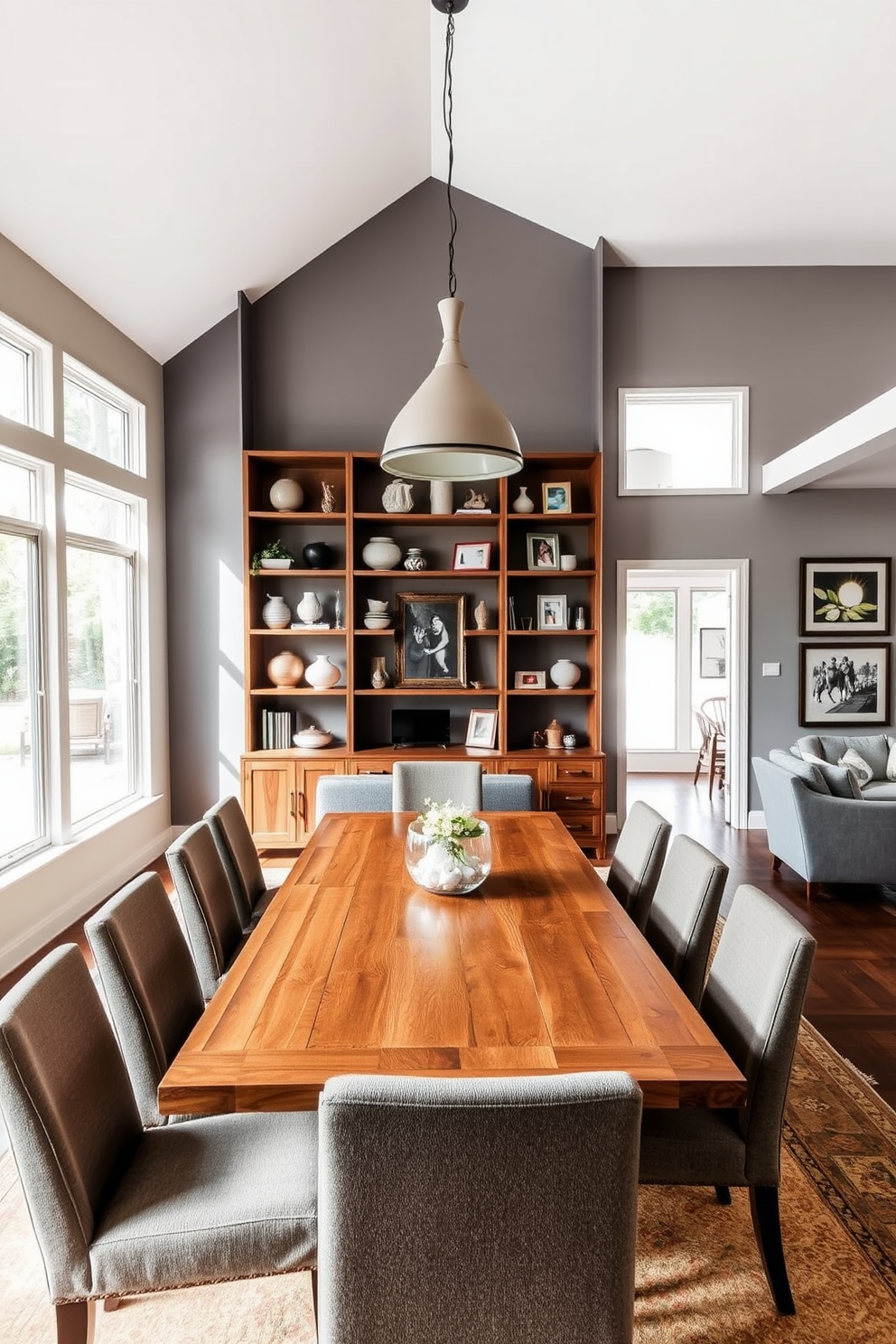 A spacious open floor plan dining room features a large wooden table surrounded by upholstered chairs. The walls are painted in a soft gray, and large windows allow natural light to flood the space, highlighting the decorative shelving along one wall. Open shelving displays an array of curated decorative items, including ceramic vases and framed artwork. A stylish pendant light hangs above the dining table, creating an inviting atmosphere for family gatherings and entertaining guests.