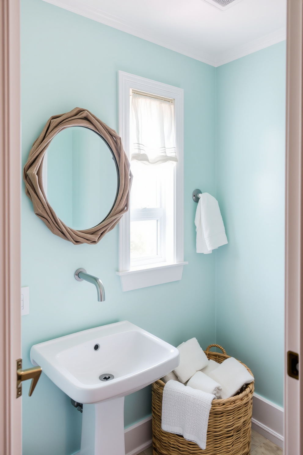 A serene powder room featuring pale aqua walls that evoke a calming beachy atmosphere. The space includes a sleek white pedestal sink and a round mirror with a driftwood frame, enhancing the coastal aesthetic. Natural light filters through a small window adorned with sheer white curtains. A woven basket filled with soft towels sits beside the sink, adding texture and warmth to the design.