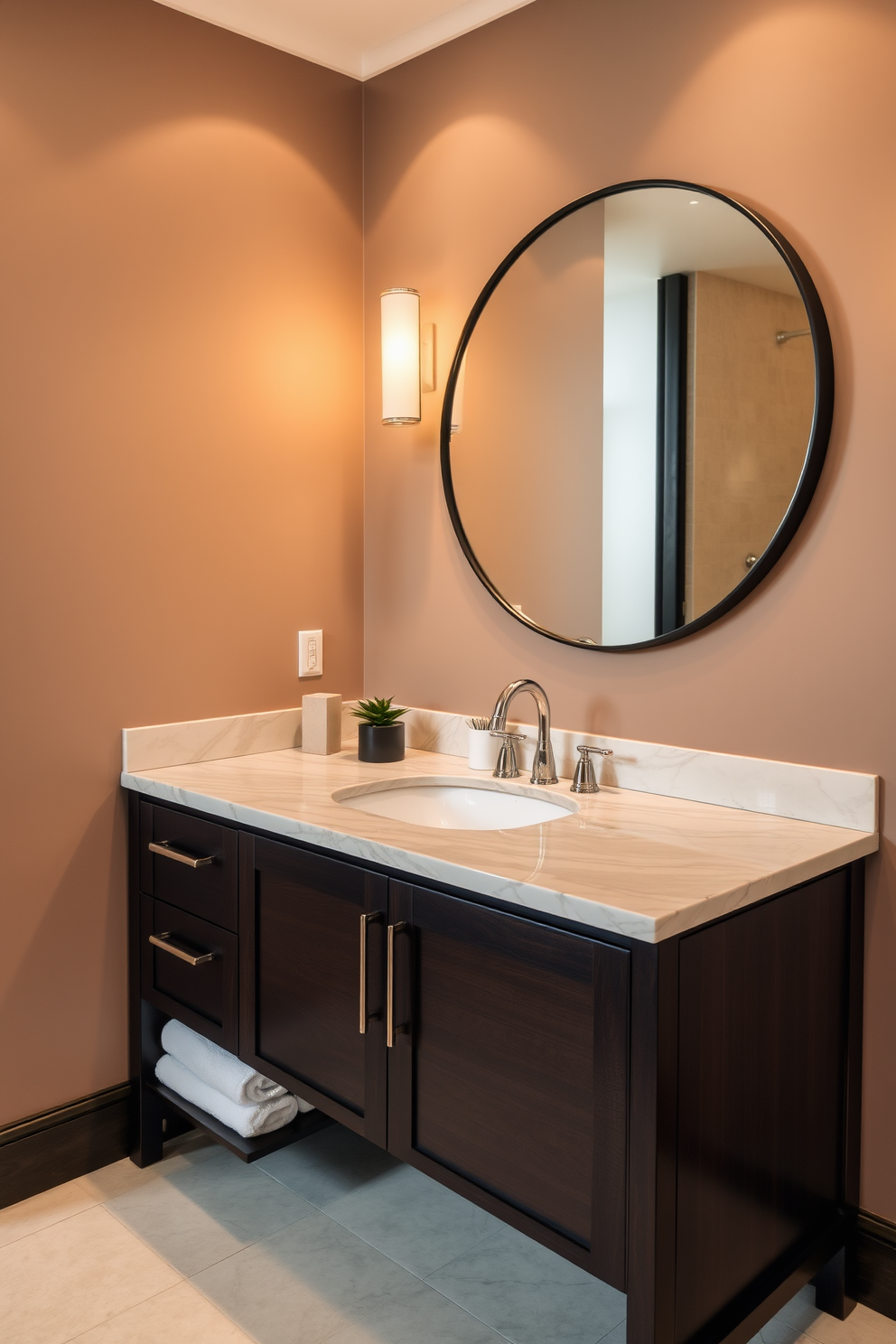 Sophisticated dark wood vanity with a polished marble countertop featuring a single elegant sink. Above the vanity, a large round mirror with a sleek black frame is mounted on the wall, reflecting the warm ambient lighting. The walls are painted in a soft taupe color, creating a cozy atmosphere. Decorative elements include a small potted plant on the countertop and luxurious hand towels neatly arranged beside the sink.