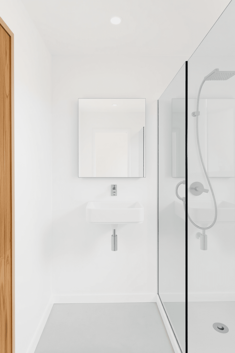 A minimalist bathroom featuring clean lines and a serene atmosphere. The space includes a wall-mounted sink with a sleek, floating vanity and a large frameless mirror above it. The walls are painted in a crisp white, creating a bright and airy feel. A simple glass shower enclosure with a rainfall showerhead complements the overall design, while natural wood accents add warmth to the space.