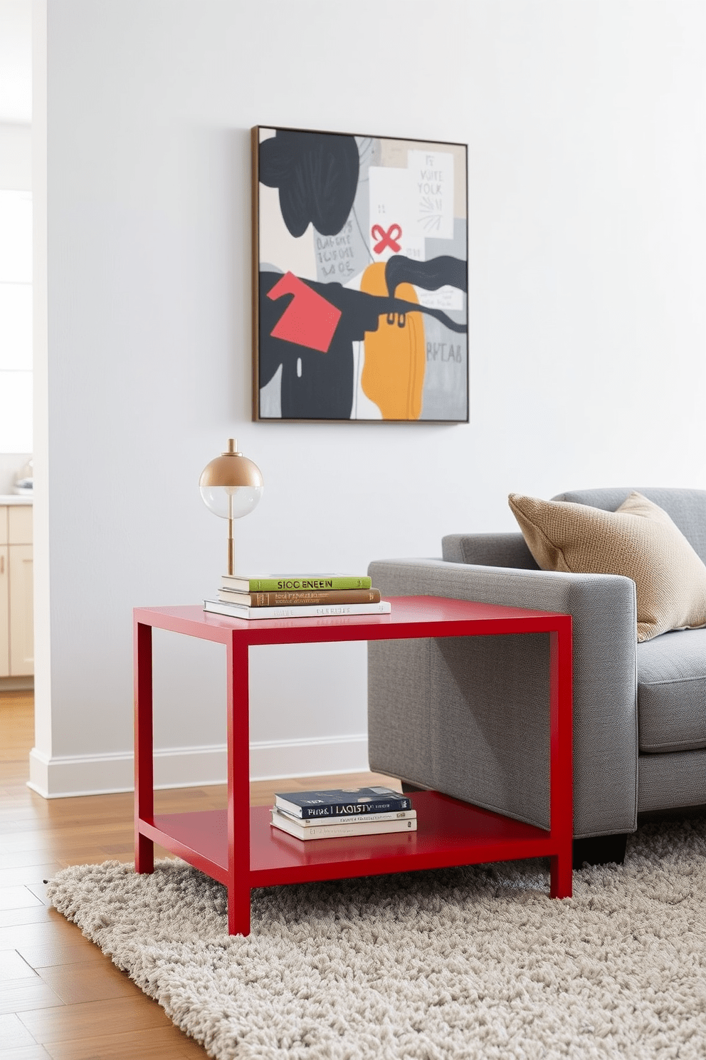 A vibrant cherry red accent table stands out in the modern living room. It is paired with a sleek gray sofa and an abstract art piece hanging on the wall. The table is adorned with a stylish lamp and a small stack of colorful books. A plush area rug in neutral tones anchors the space, adding warmth and texture.