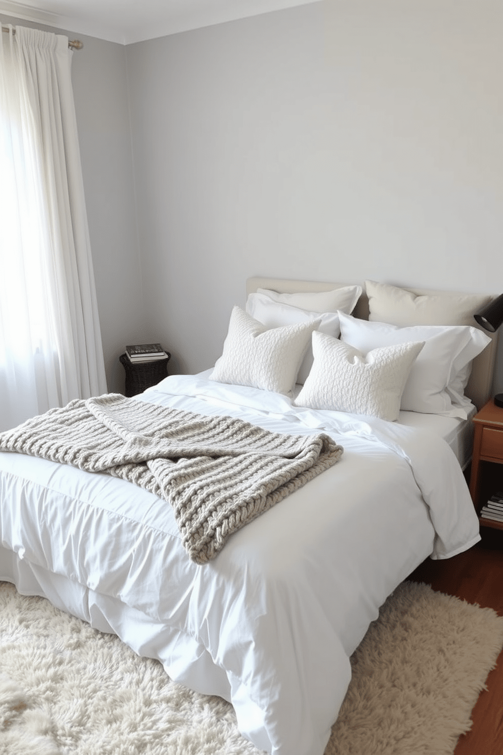 A cozy bedroom featuring a soft bed dressed in white linens topped with an array of textured pillows and a chunky knit throw. The walls are painted in a calming light gray, and a wooden nightstand holds a small lamp and a stack of books for a personal touch. The floor is adorned with a plush area rug that adds warmth to the space. Natural light filters in through sheer curtains, creating a serene and inviting atmosphere.