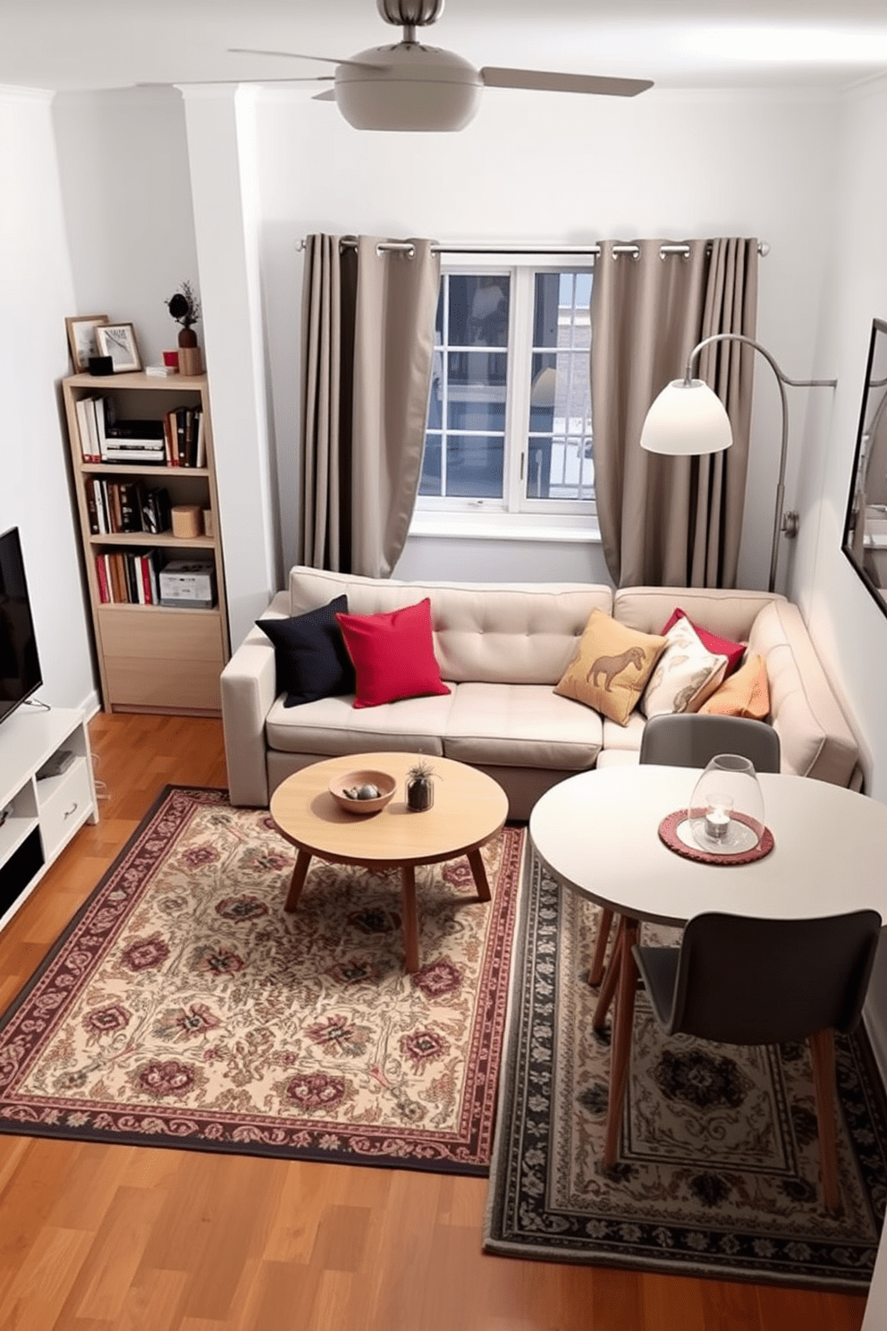 A cozy small apartment living room featuring a light gray sofa adorned with colorful throw pillows. A round coffee table sits in the center, surrounded by a patterned area rug that defines the seating area. To the left, a compact bookshelf filled with books and decorative items adds personality to the space. A second area rug beneath a small dining table creates a distinct dining zone, enhancing the functionality of the room.