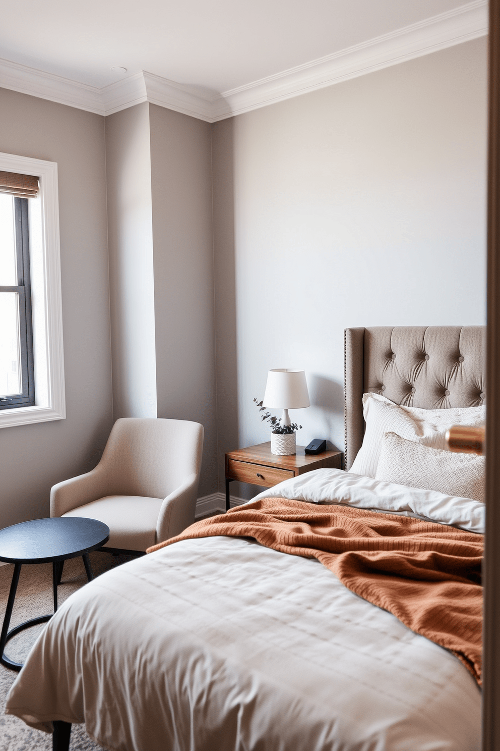 A cozy small bedroom featuring a comfortable bed with a plush headboard positioned against a soft gray wall. To the left of the bed, there's a small seating area with a stylish armchair and a round side table, perfect for reading or relaxing. The bedding is layered with warm textures and neutral colors, creating an inviting atmosphere. A minimalist desk is placed near the window, adorned with a small potted plant to bring a touch of nature indoors.