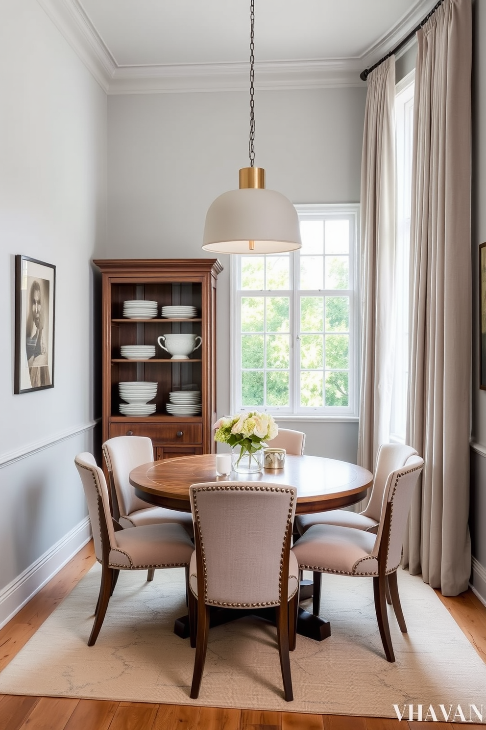 A cozy small dining room featuring a round wooden table surrounded by upholstered chairs in soft pastel colors. The walls are adorned with light gray paint, and a tall cabinet with open shelving showcases elegant dishware and decorative items. A statement pendant light hangs above the table, adding warmth and character to the space. Floor-to-ceiling curtains frame a window, allowing natural light to fill the room while providing a sense of height and openness.