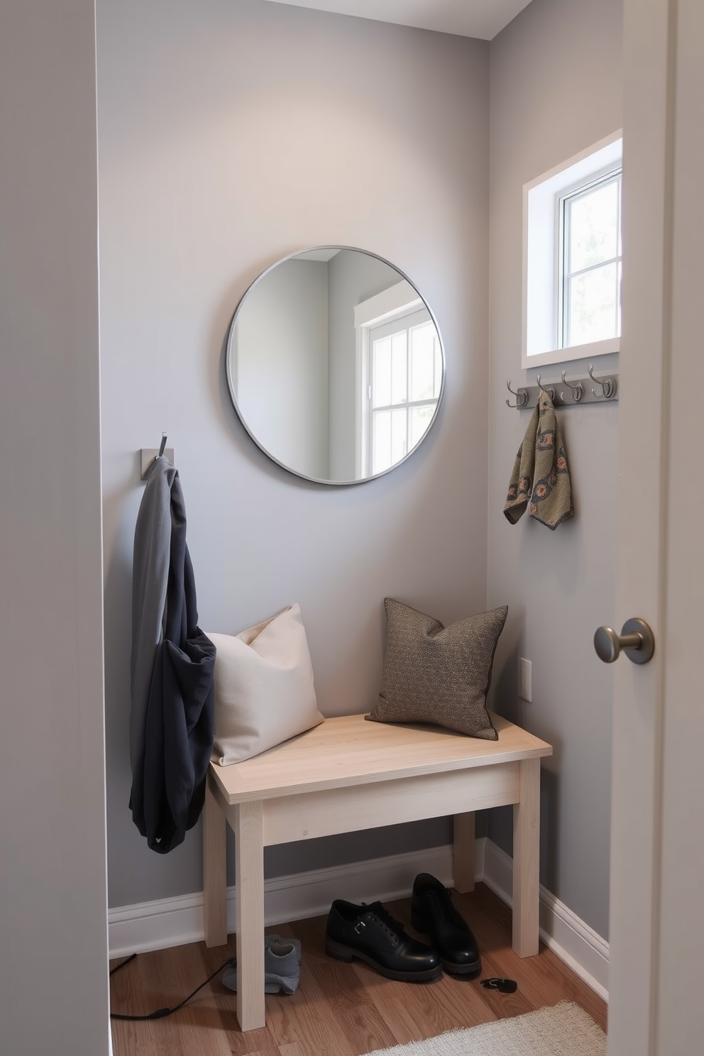 A small entryway features wall-mounted coat hooks arranged neatly along one side. The hooks are made of brushed nickel and complement a light wood bench that provides a place to sit while putting on shoes. The walls are painted in a soft gray hue, enhancing the sense of space. A round mirror hangs above the bench, reflecting natural light and making the area feel more open and inviting.